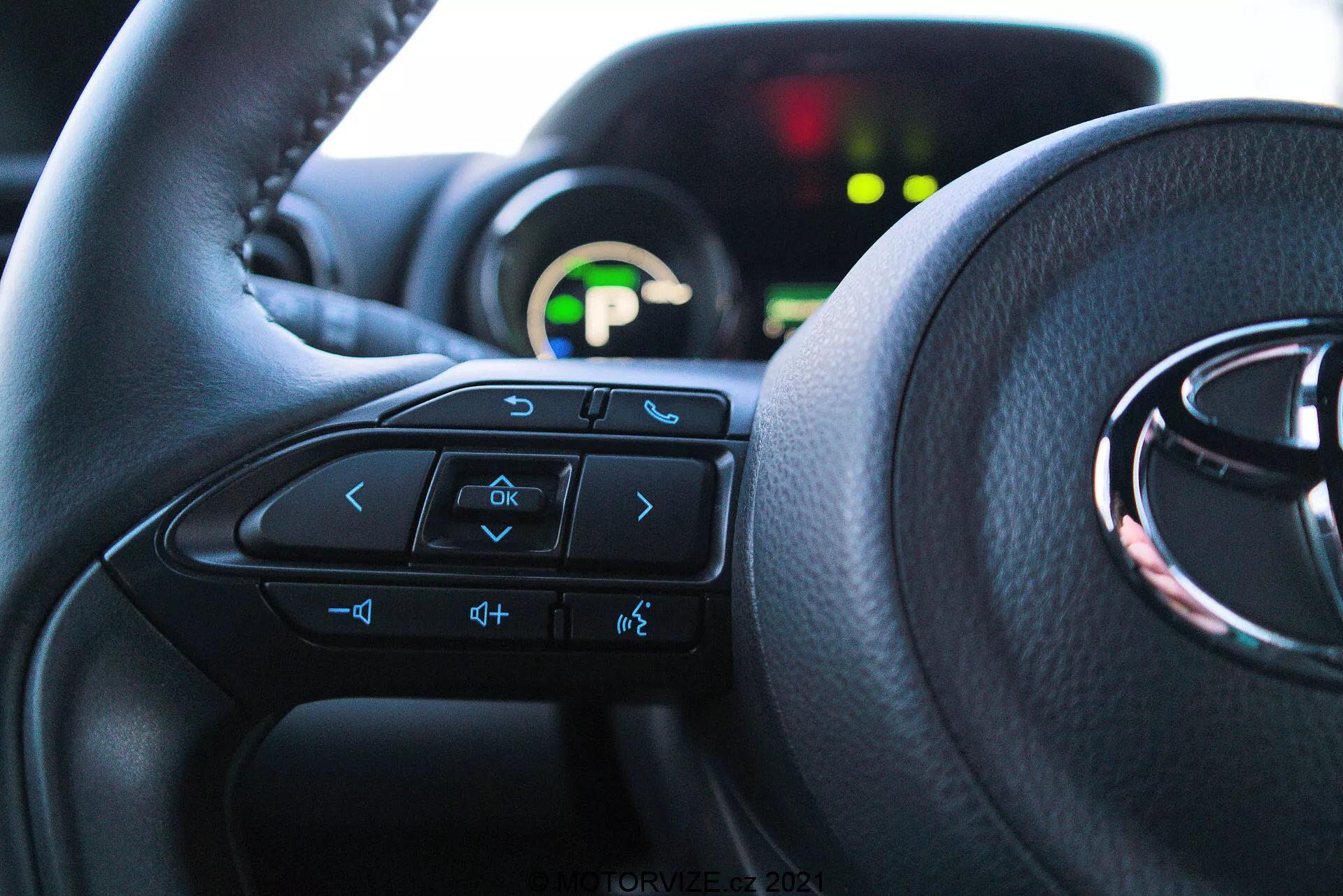 Närbild av en interiör i en Toyota Yaris 2020 från förarsätets perspektiv, med fokus på ratten med multifunktionsknappar och ett instrumentkluster i bakgrunden. Ratten dominerar de nedre två tredjedelarna av bilden, med pilknappar, röstkommandon, volymkontroller och underhållningssystemknappar synliga. Det delvis synliga instrumentklustret är belyst med en grön digital display och är oskarpt på grund av ett grunt skärpedjup. Naturligt ljus lyser upp interiören och betonar komfort och ett användarvänligt gränssnitt.