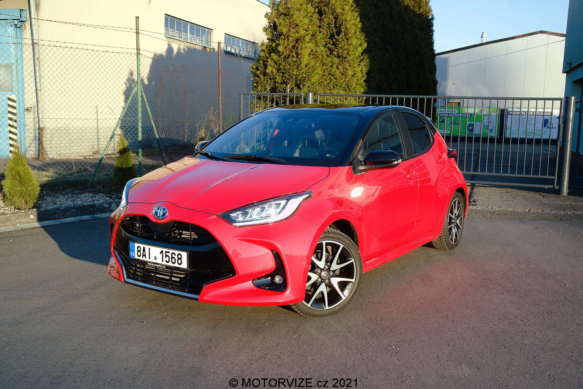 Bild av en ljusröd 2020 Toyota Yaris halvkombi parkerad i vinkel, som visar både front- och sidoprofiler. Strålkastardesignen är skarp och kantig, grillen har ett honungsmönster med en centralt placerad emblem, och den främre stötfångaren har synliga konturer med dimljus. Sidprofilen avslöjar tvåfärgade lättmetallfälgar, färgmatchade dörrhandtag och speglar med skarpa linjer och konturer längs sidan. Den blanka lacken antyder ett nytt eller väl underhållet tillstånd. Bakgrunden inkluderar en klar himmel, industriella strukturer och mjuka skuggor som indikerar en utomhusmiljö, troligen en parkeringsplats nära en kommersiell eller industriell anläggning.