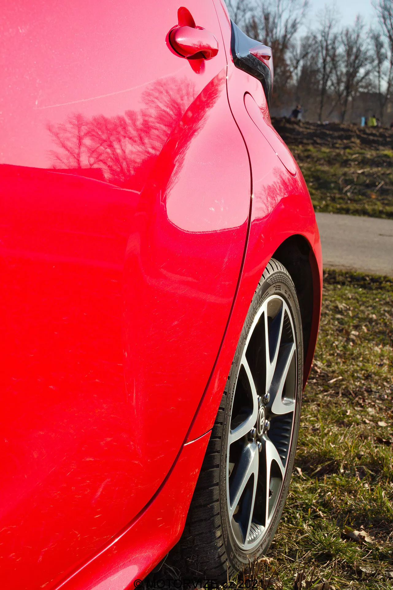 Närbild av sidan på en röd 2020 Toyota Yaris bakre kvartspanel tagen i något uppåtvinklat perspektiv som framhäver den ljusröda färgen, den multiplicerade fälgen, däckmönstret, den rena kurvan av den bakre kvartspanelen och stötfångaren, glansig lackreflektion med träd och en klarblå himmel samt synligt bränslelock och trimdetalj.
