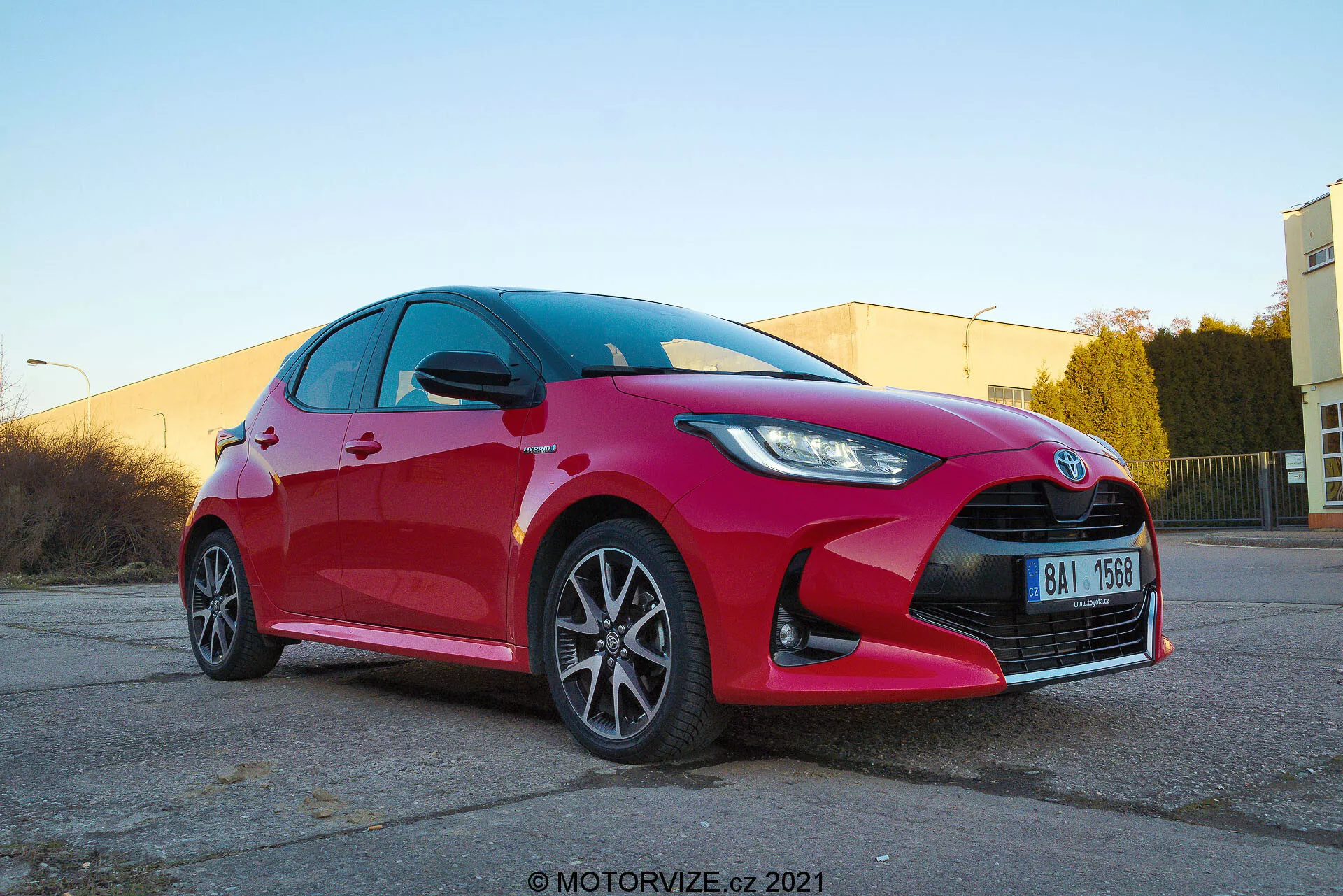 Lågvinklad vy av en modern Toyota Yaris hatchback 2020 i livfull röd färg, fotograferad framifrån i trekvartsperspektiv som visar skarpa och kantiga LED- eller HID-strålkastare, framträdande galler med centralt emblem, flerspoks lättmetallfälgar, integrerade blinkers i sidospeglarna, rena reflekterande ytor och möjligen prestandainriktat bromssystem. Bilden togs i ett varmt upplyst industriellt parkeringsområde med vattenstämpel motorvize.cz 2021.