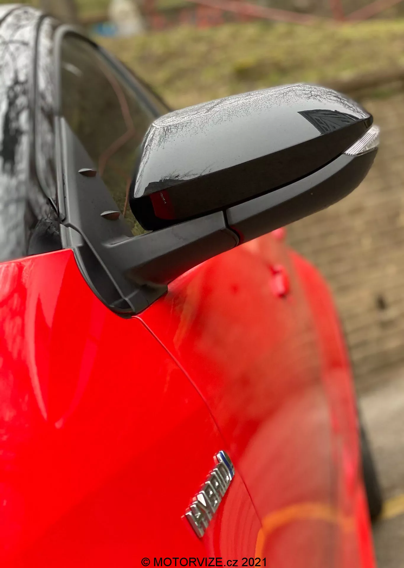 Närbild av en utdragen sidospegel på en Toyota Yaris 2020, med ett elegant, aerodynamiskt svart hölje som kontrasterar med bilens glansiga röda kaross. Spegeln betonar tekniska inslag som möjlig elektrisk justering, uppvärmning och integrerade blinkers, även om specifika funktioner inte är bekräftade. Vattendroppar antyder nyligen regn eller en biltvätt, medan en suddig bakgrund och ett grunt skärpedjup håller fokus på spegeln. En del av en badge på framskärmen är synlig men suddig.