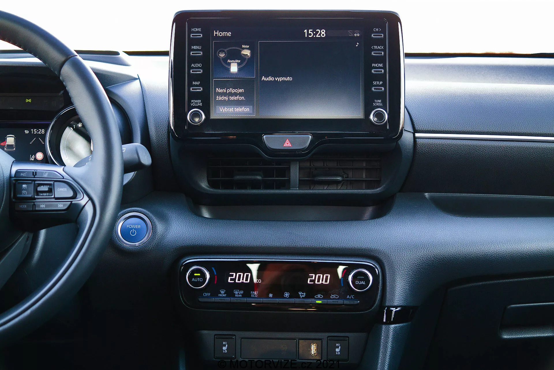 Närbild av den centrala instrumentbrädan på en Toyota Yaris 2020 från ett inre perspektiv. Ratten är delvis synlig till vänster, vilket indikerar en synvinkel från passagerarsätet eller centralt i bilen. Instrumentbrädan har en framträdande infotainment-pekskärm längst upp på mittkonsolen, som visar en hemskärm med olika ikoner och tiden '15:28'. Nedanför finns en digital klimatkontrolldisplay med inställningar för tvåzonstemperatur, båda sidor inställda på '20.0' grader Celsius. Ytterligare kontroller för avfrostning och möjligen förarassistanssystem är synliga under klimatkontrollerna. Ovanför infotainment-skärmen finns två horisontella luftventiler med en varningsljusbrytare mellan dem. En start/stopp-knapp är delvis synlig längst till vänster, vilket indikerar nyckellös tändningsteknologi. En högtalargaller finns i det övre vänstra hörnet. Instrumentbrädans material har både släta och texturerade element, vilket tyder på fokus på komfort och teknologi.