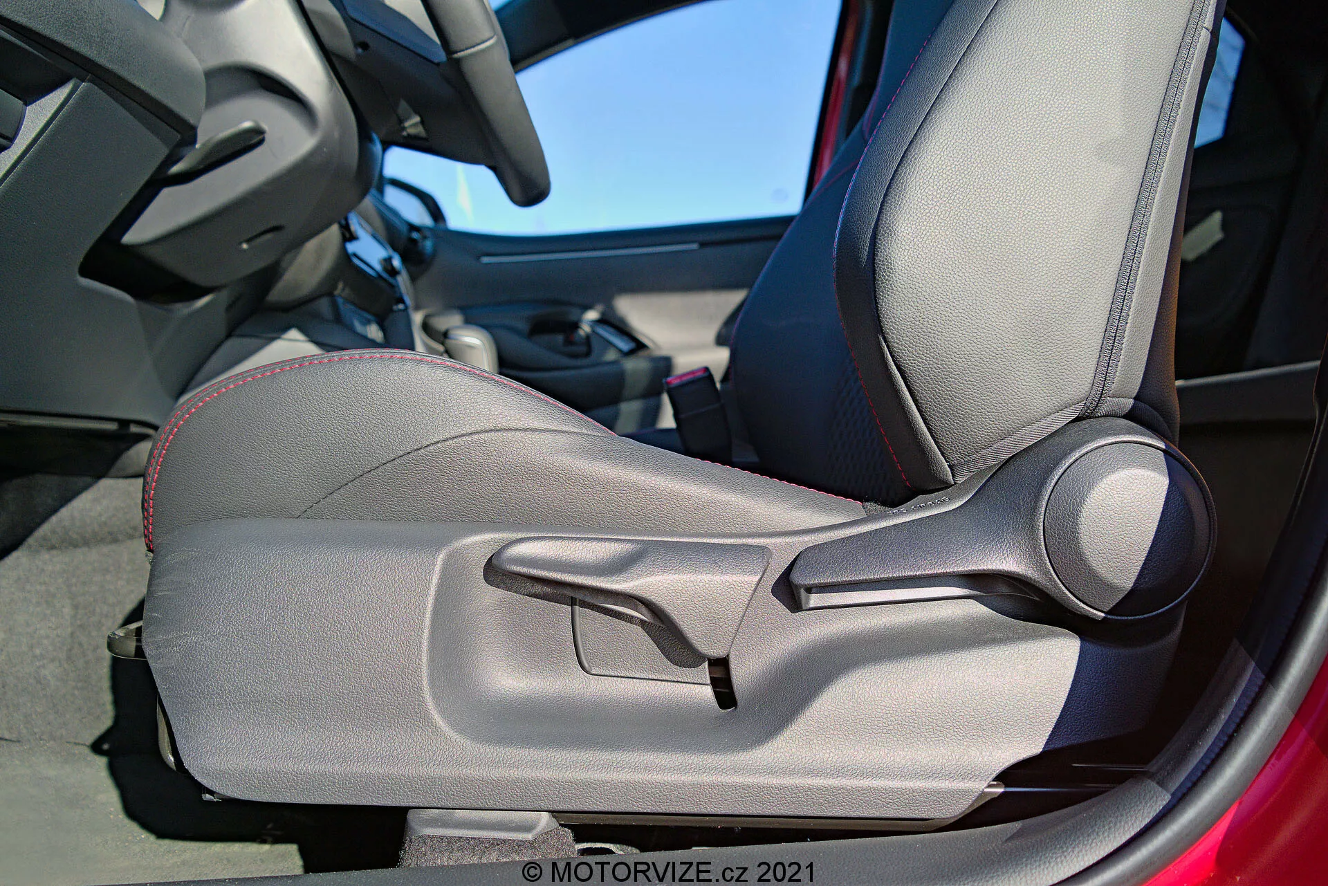 Intern detaljbild av 2020 Toyota Yaris som fokuserar på förarsidans dörr och säte, med betoning på tvåtonat tyg, dekorativa sömmar, konturerad design för ergonomiskt stöd, manuellt justerbara sidostöd och en framträdande manuell sätesjusteringsspak. Dörrkarmen är delvis synlig och antyder att dörren är öppen.