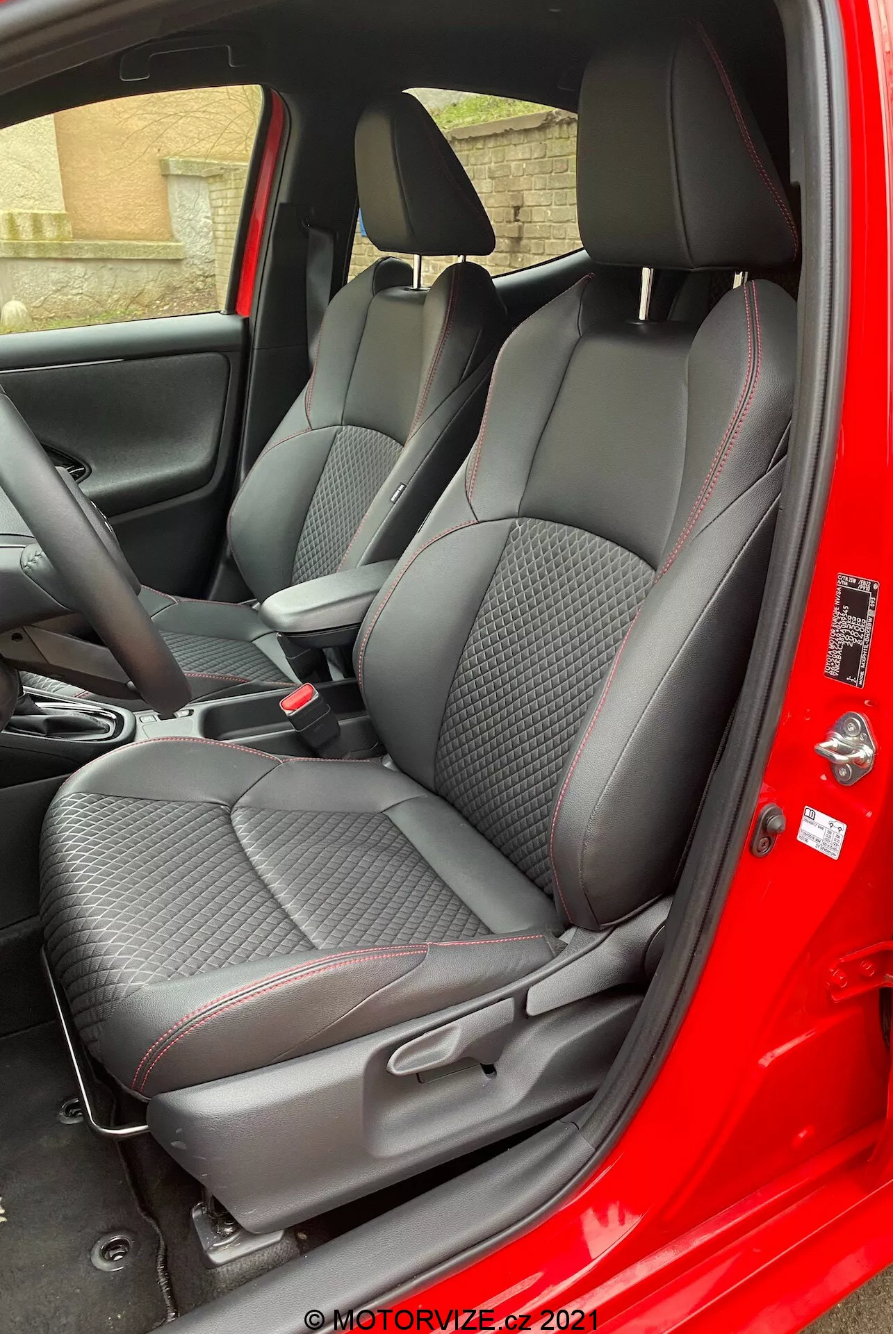 Interiören av 2020 Toyota Yaris sedd från passagerarsidan med dörren öppen, som framhäver premiumtexturerade tygsäten med diamantmönstrad sömnad och röda accenter, justerbara nackstöd, centralkonsolens kontroller och dörrpanel med kolfiberliknande textur samt elektriska fönsterkontroller.