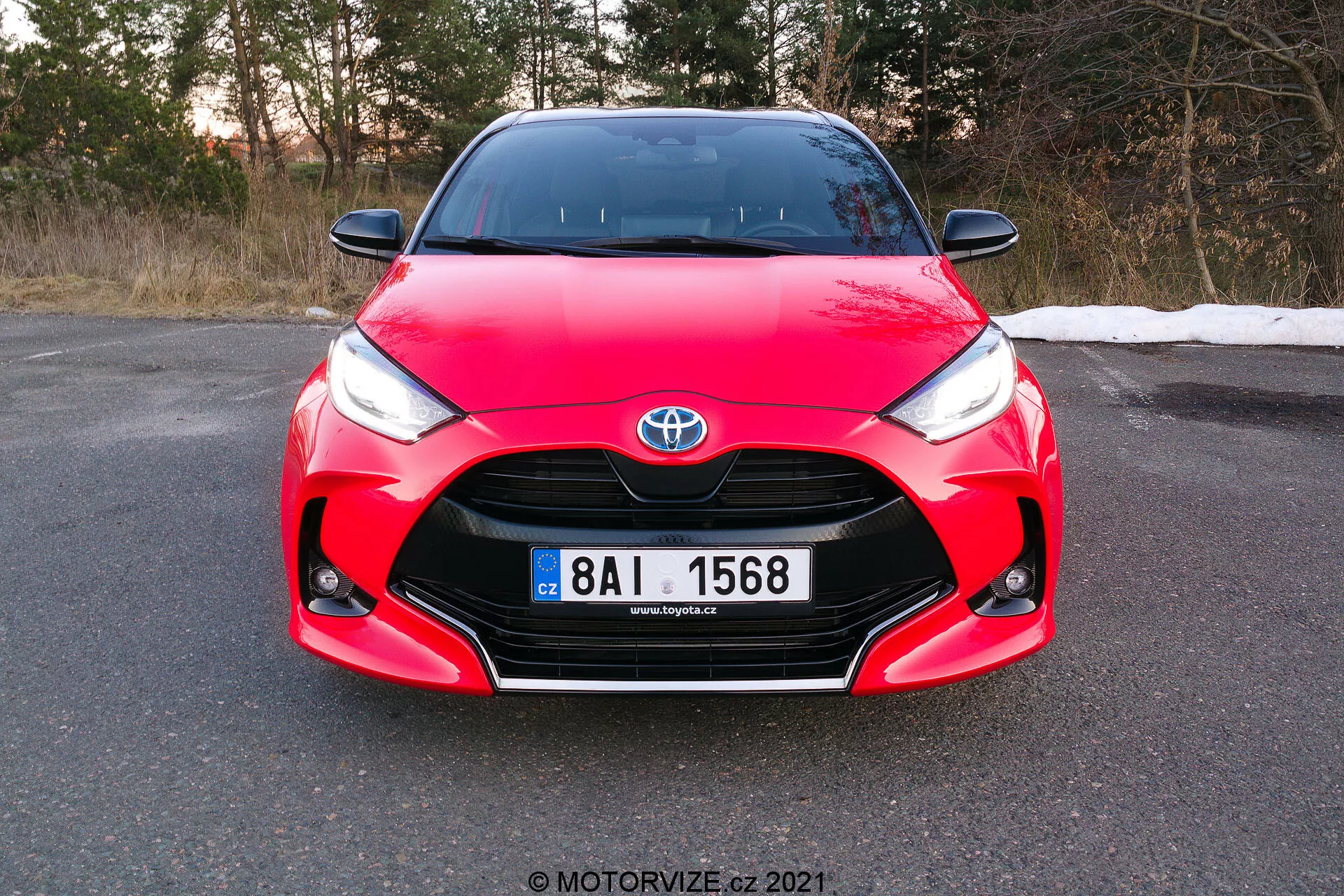 Bilden visar en frontal vy av en röd 2020 Toyota Yaris, tagen i en lätt vinkel med fokus på frontgrillen, strålkastarna och motorhuvsområdet, med registreringsskylten helt synlig. Bilen har distinkta designelement inklusive Toyota-emblemet centrerat på grillen, skarpa vinklade strålkastare som smalnar av mot grillen och komplexa kurvor på motorhuven, vilket visar dess aerodynamiska och sportiga design. Fotot, taget under dagen före skymningen, antyder att bilen är parkerad på en plan yta i en miljö som kan vara sen höst eller tidig vår.