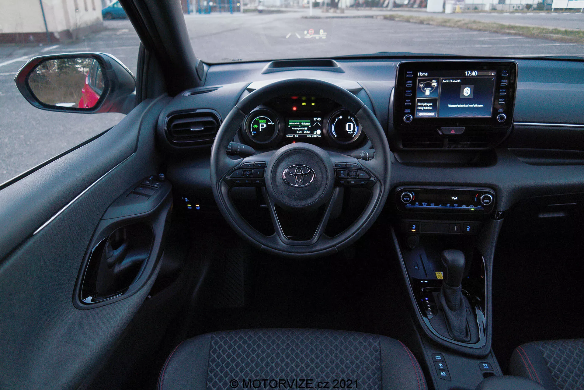 Interiörperspektiv från förarsätet i en Toyota Yaris 2020, som visar ratt med inbyggda reglage, digitalt instrumentkluster och en infotainmentskärm. Interiören har klimatanläggningsknappar, en växelväljare och ett övervägande svart färgschema med texturerade ytor och kromade detaljer.