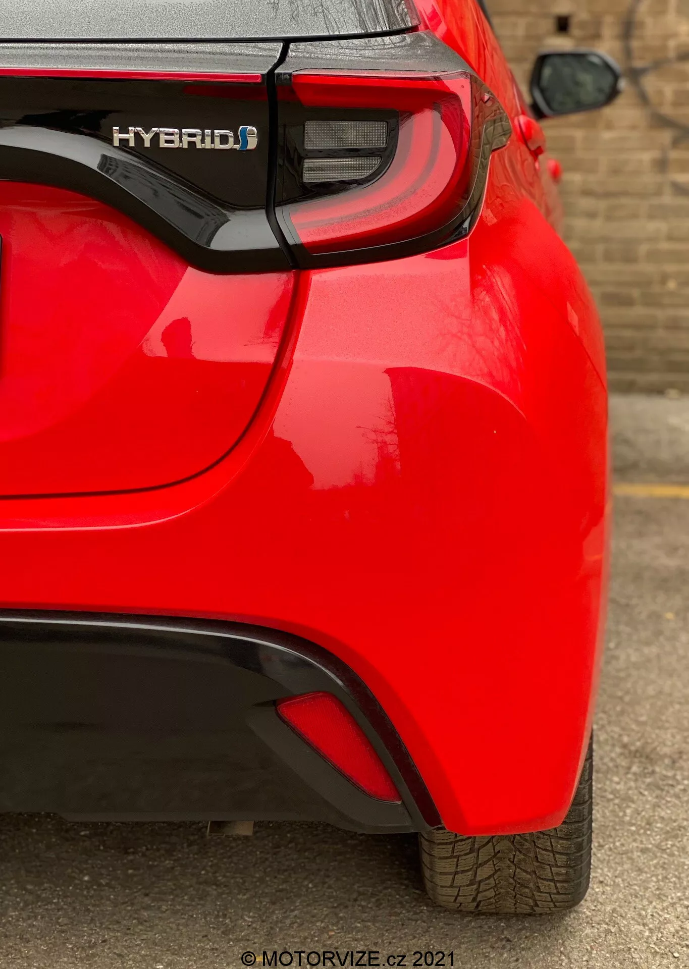 Närbild av bakifrån av en röd Toyota Yaris hybrid 2020 med fokus på höger baksida. Fångar funktioner som 'HYBRID' märkningen, modern LED-baklyktkluster, subtil aerodynamisk böj på den bakre stötfångaren och en synlig parkeringssensor. Reflekterande, glansig lack och välunderhållet däckmönster tyder på ett nytt eller välvårdat fordon.