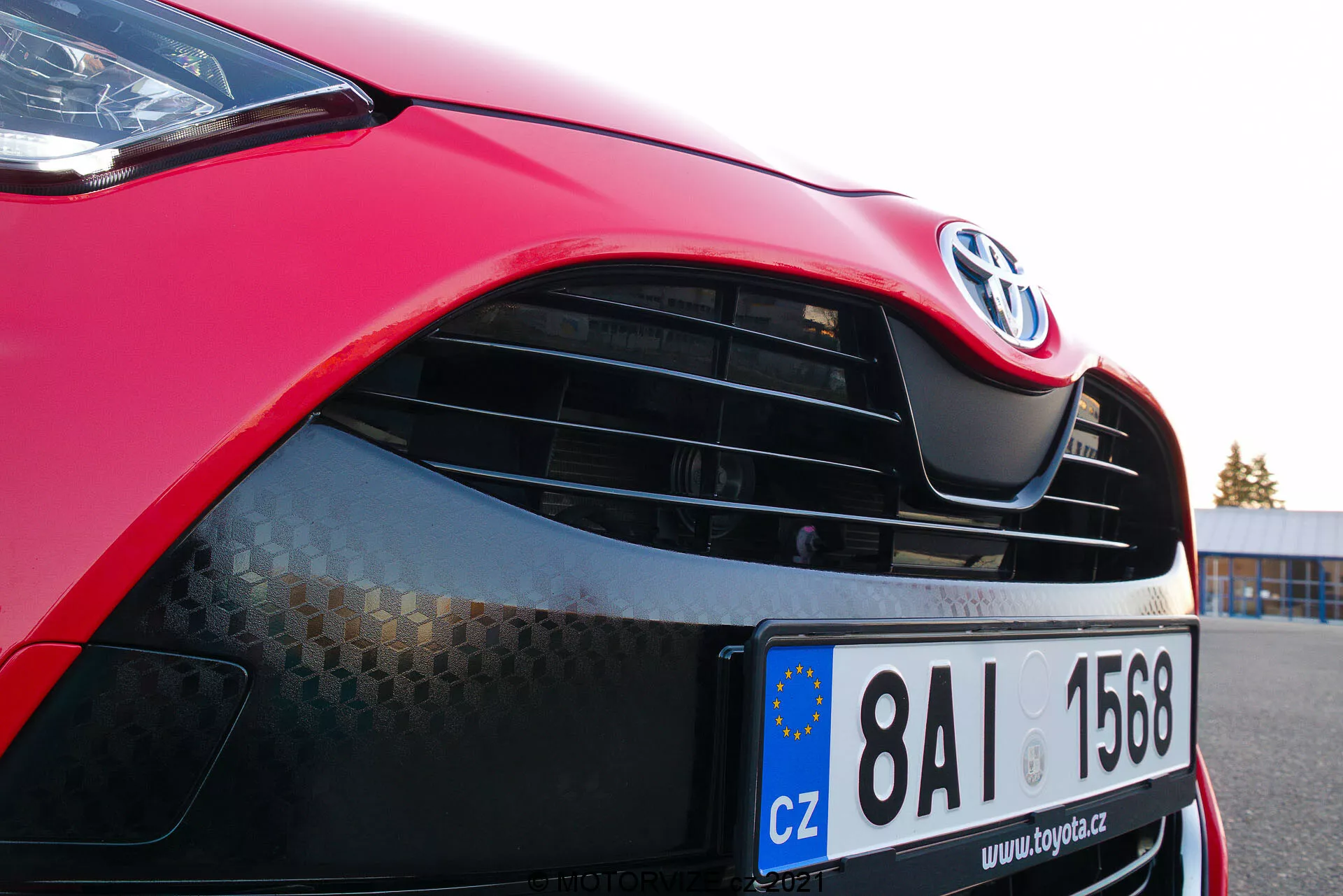Närbild av en röd 2020 Toyota Yaris som fokuserar på bilens vänstra framsida, med motorhuven, den moderna strålkastaren med möjlig LED- eller HID-teknologi, grillen med Toyota-emblem och honungsmönster, glansig röd färg och delvis synlig främre stötfångare med lägre luftintag. EU-registreringsskylt antyder registrering i Tjeckien.