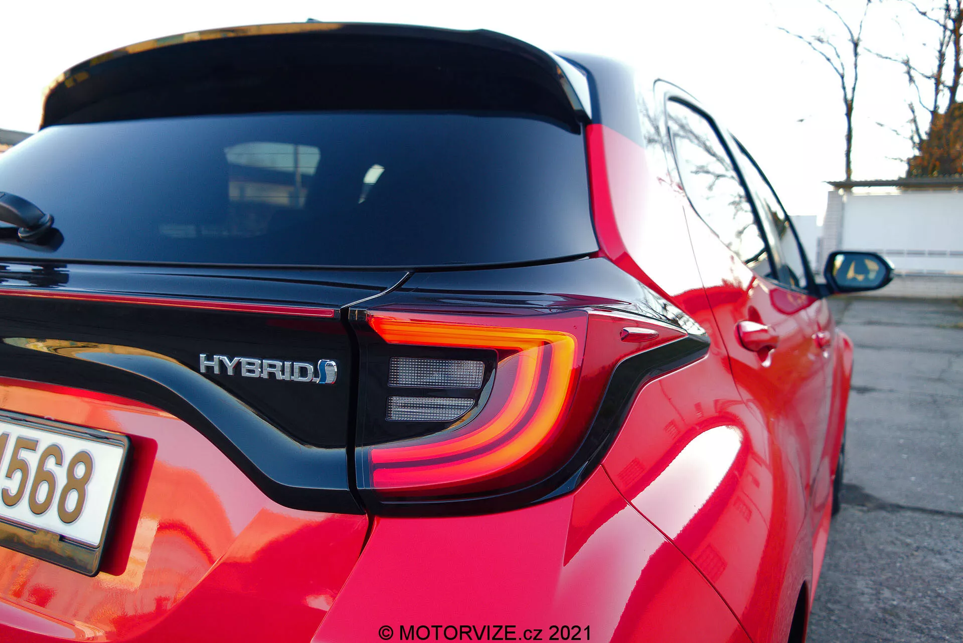 Närbild, vinklad bild av bakre delen av en röd Toyota Yaris 2020, som framhäver den moderna LED-baklyktan med kurviga linjer, HYBRID-märket, integrerad reflex i bakre stötfångaren, bakre vindrutetorkaren, takspoiler, avfrostningslinjer i bakrutan, reflekterande röd lack och delvis oskarpt nummerplåtsområde med backkamera.