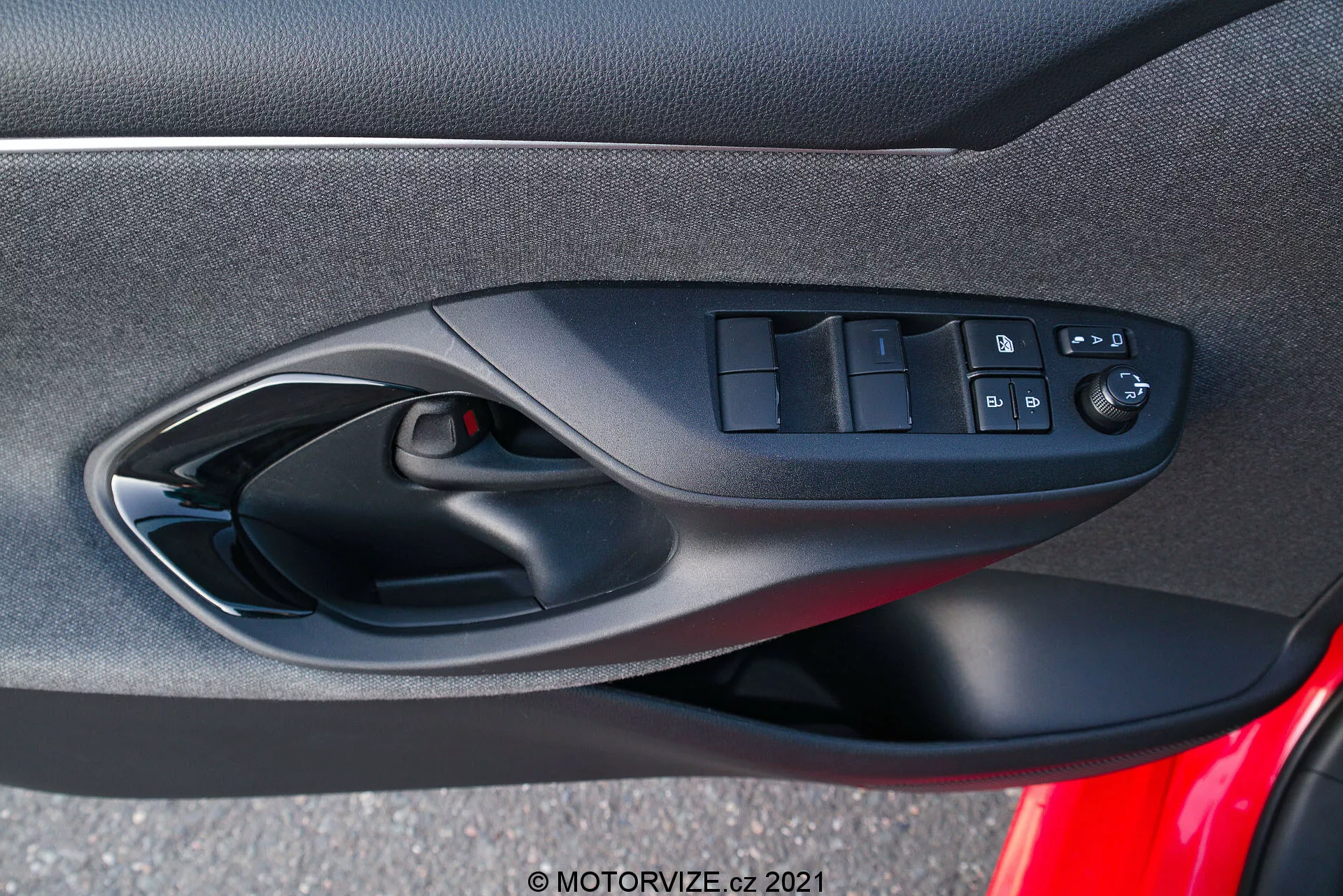 Interiördörrpanel av en Toyota Yaris 2020 som fokuserar på de ergonomiskt utformade förarsidadörrkontrollerna, inklusive strömbrytare för elhissar, kraftdörrlåsknappen och elektroniskt justerbara spegelkontroller, fångad från en passagerares perspektiv inne i fordonet. Fotot framhäver blandningen av mjuka tyg- eller komposita material, det svarta färgschemat och ett synligt högtalargaller som är en del av bilens ljudsystem.