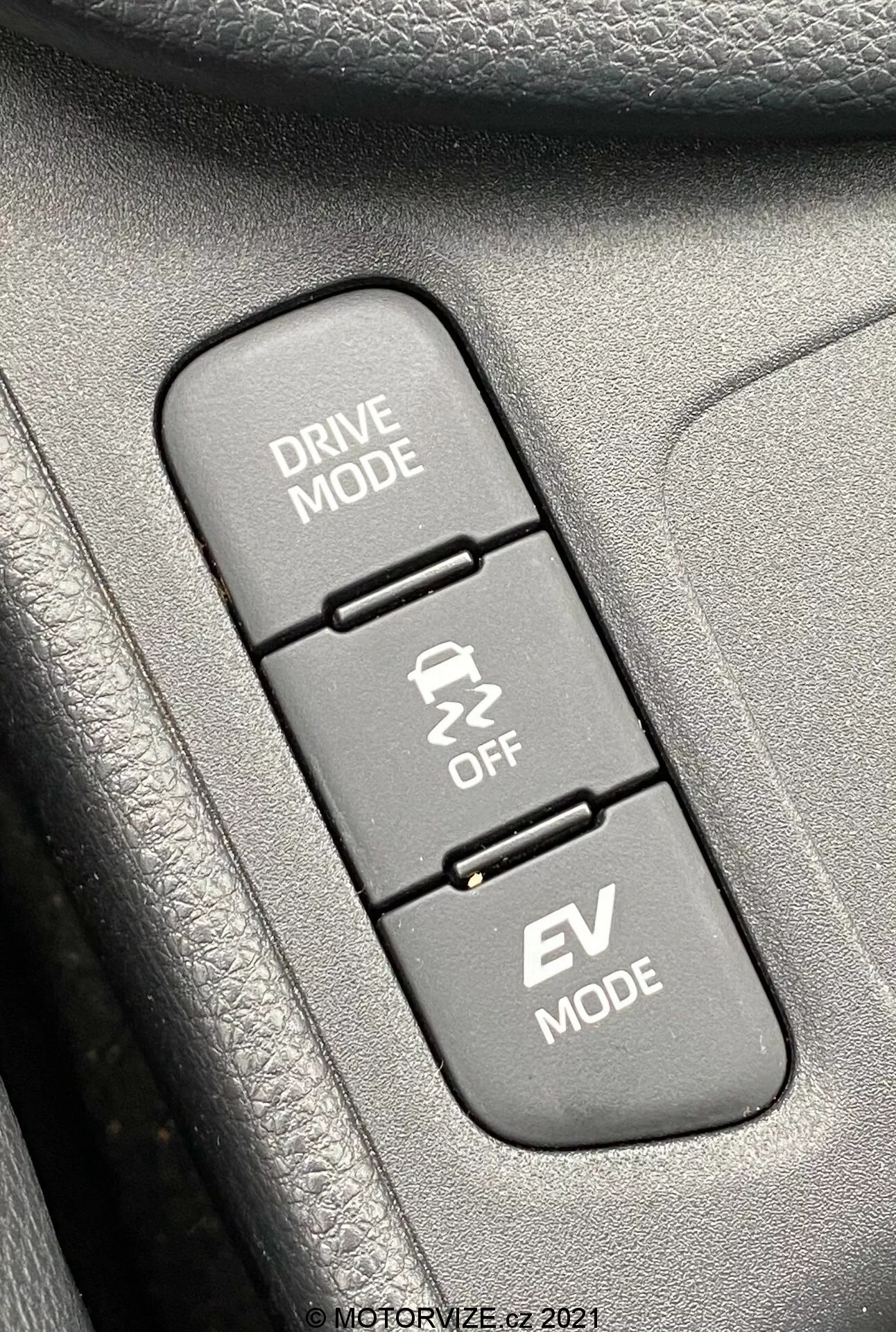 Närbild av interiören i en Toyota Yaris 2020, med fokus på tre vertikalt uppradade kontrollknappar relaterade till körlägen. Uppifrån och ner: 'DRIVE MODE' med en ikon av en slingrande väg, 'Traction Control OFF' med en ikon av en sladdande bil och 'EV MODE' som indikerar elbilsläge. Knapparna är monterade på en texturerad yta, troligen en del av en dörrpanel eller mittkonsol. Bilden är skarpast på knapparna och de omgivande texturerna är något suddiga, upplyst av dagsljus för klar synlighet.