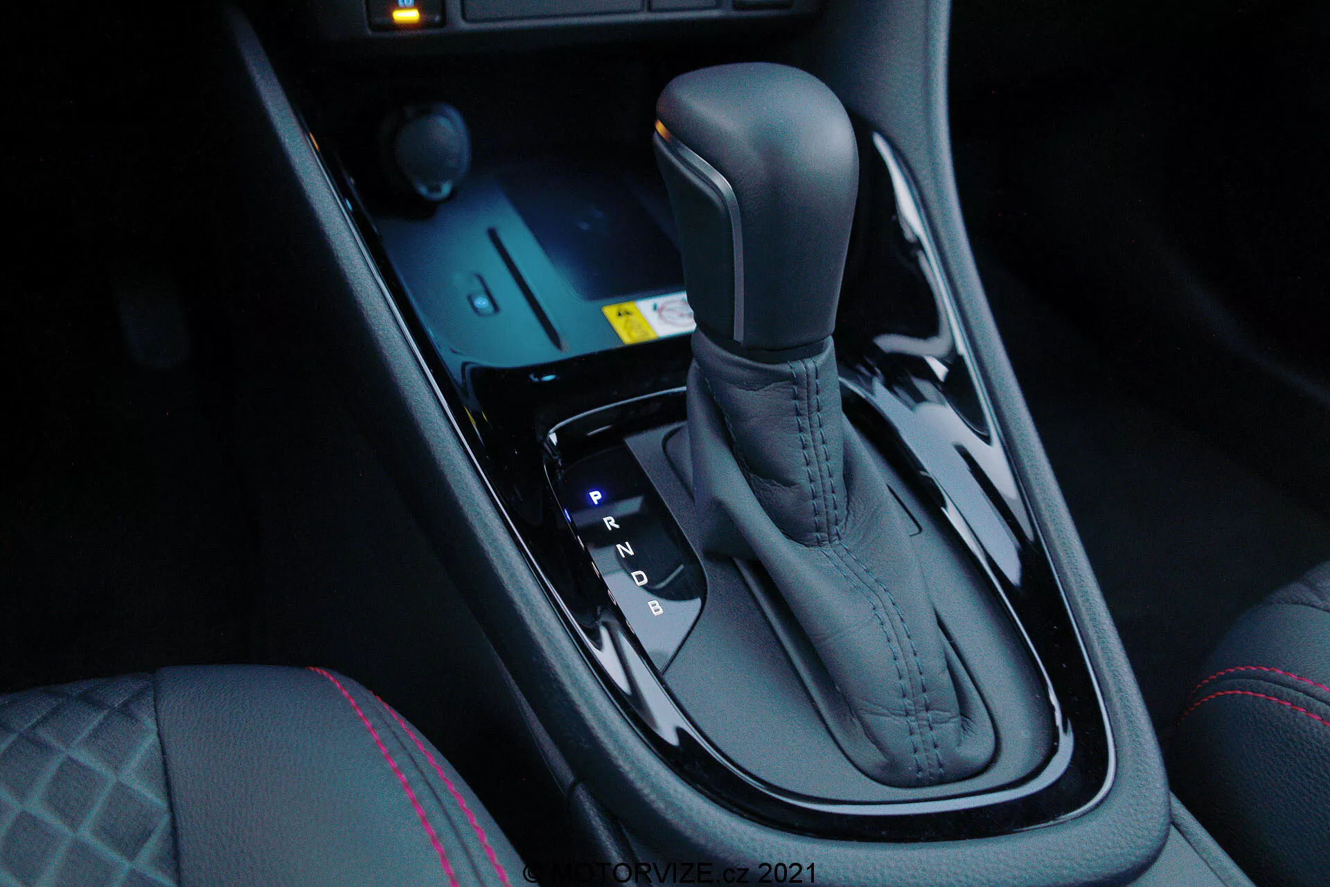 Närbild av Toyota Yaris 2020 automatiska växelväljare, sett från framsätespassagerarens sida i en nedåtvinklad position, som lyfter fram den eleganta metalliska finishen och ergonomiska designen. Växelväljaren är centralt placerad mellan förar- och passagerarsätena och innefattar positionslägena Park, Reverse, Neutral och Drive, samt manual växlingsmöjlighet (+ och -). Framför växelväljaren finns elektroniska kontroller för körlägen och traction control, tillsammans med ett blått upplyst startknappssystem. Den mörka, glansiga mittkonsolen tillför en modern lyxkänsla, kompletterad med sätesvärmekontroller och särskild sömnad på förarsätet, vilket antyder ökad komfort och avancerad kördynamik.