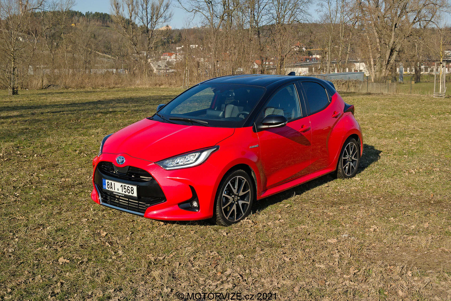 Bilden visar en livfull röd 2020 Toyota Yaris, vilande på ojämn gräsklädd terräng i ett halvlandsbygdsmiljö, sedd från ett tredjedelsperspektiv. Bilens moderna, aerodynamiska design har skarpa vinklar runt strålkastarna och en kompakt, sportig utseende. De mörka multiekerhjulen och stiliserade LED-strålkastarna är synliga. Sidofönstren och framrutan ger en mörk inblick i interiören, vilket antyder potentiella avancerade bilsystem.