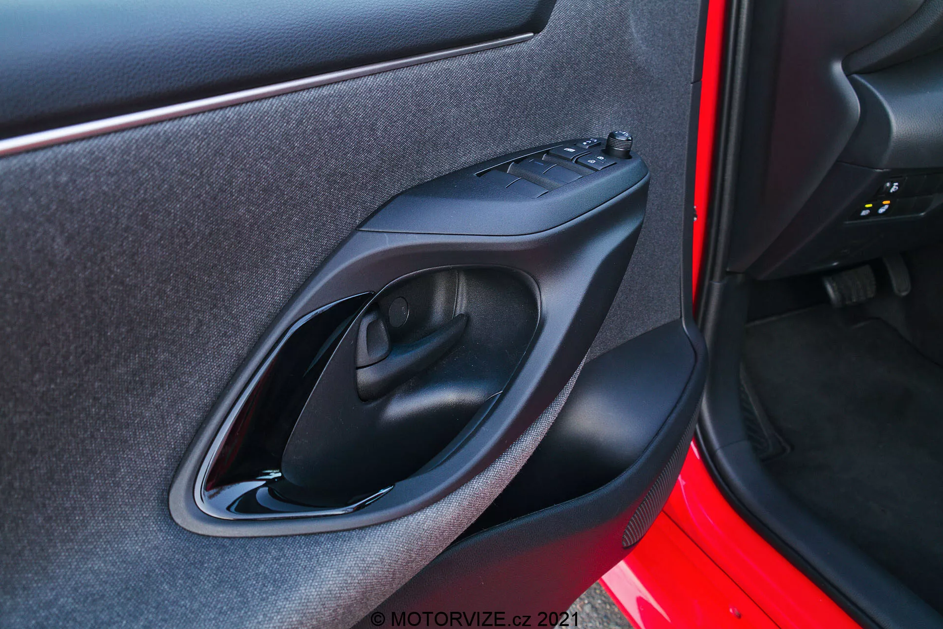 Närbild av dörrpanelen på en Toyota Yaris 2020 tagen från ett exteriört perspektiv. Bilden fokuserar på den högra dörrpanelen med design- och teknologifunktioner som ett dörrhandtag i matt svart plast, kontrollpanel för elektriska fönster, ergonomiskt utformad armstöd och en liten förvaringsficka. Dörrens utsida är målad i en livfull röd färg.