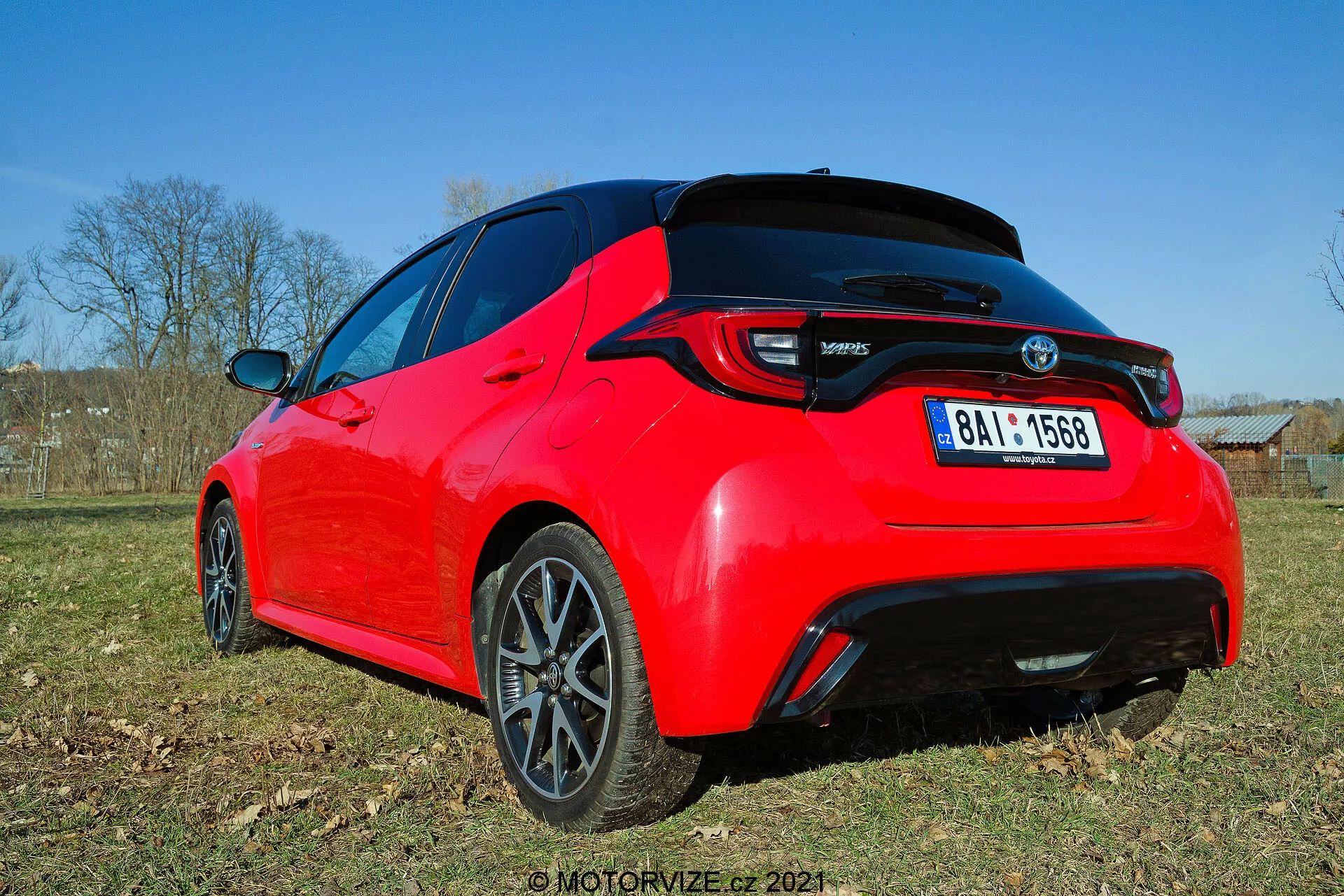 Vy av bakifrån och tre kvarts vinkel av en ljusröd Toyota Yaris 2020 hatchback, fotograferad från låg vinkel, vilket framhäver desiglinjer och nyckelfunktioner som LED-bakljus, bakspoiler, hajfensantenn och mörkfärgade flerspakiga lättmetallfälgar. Bilden fångar detaljen av bakdelen och delvis sidprofilen i naturligt dagsljus, vilket visar upp dess sportiga estetik och moderna element.