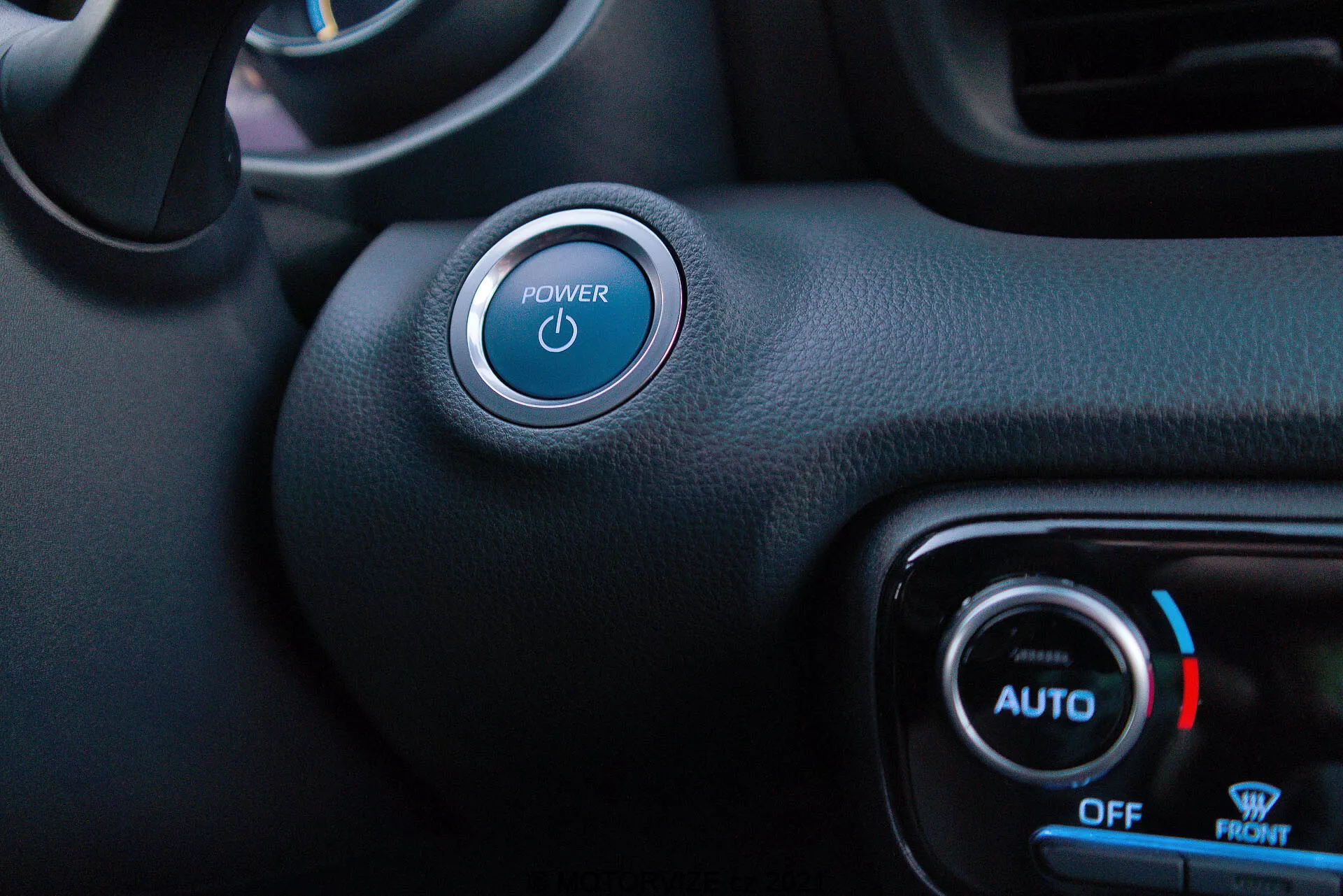 Inre vy från förarsidan på en Toyota Yaris 2020 som framhäver Start/Stop-knappen och klimatkontrollen. Strömknappen med en blåt upplyst symbol är framträdande i det övre vänstra hörnet med en silverbeklädnad. Nedanför och till höger syns klimatkontrollvred med blått ljus och en snöflingesymbol för kyltemperatur, med etiketter 'AUTO', 'OFF' och 'FRONT'. En mix av texturerad och slät svart syntetisk material visar på en minimalistisk stil med funktioner som nyckellös start och automatisk klimatkontroll.