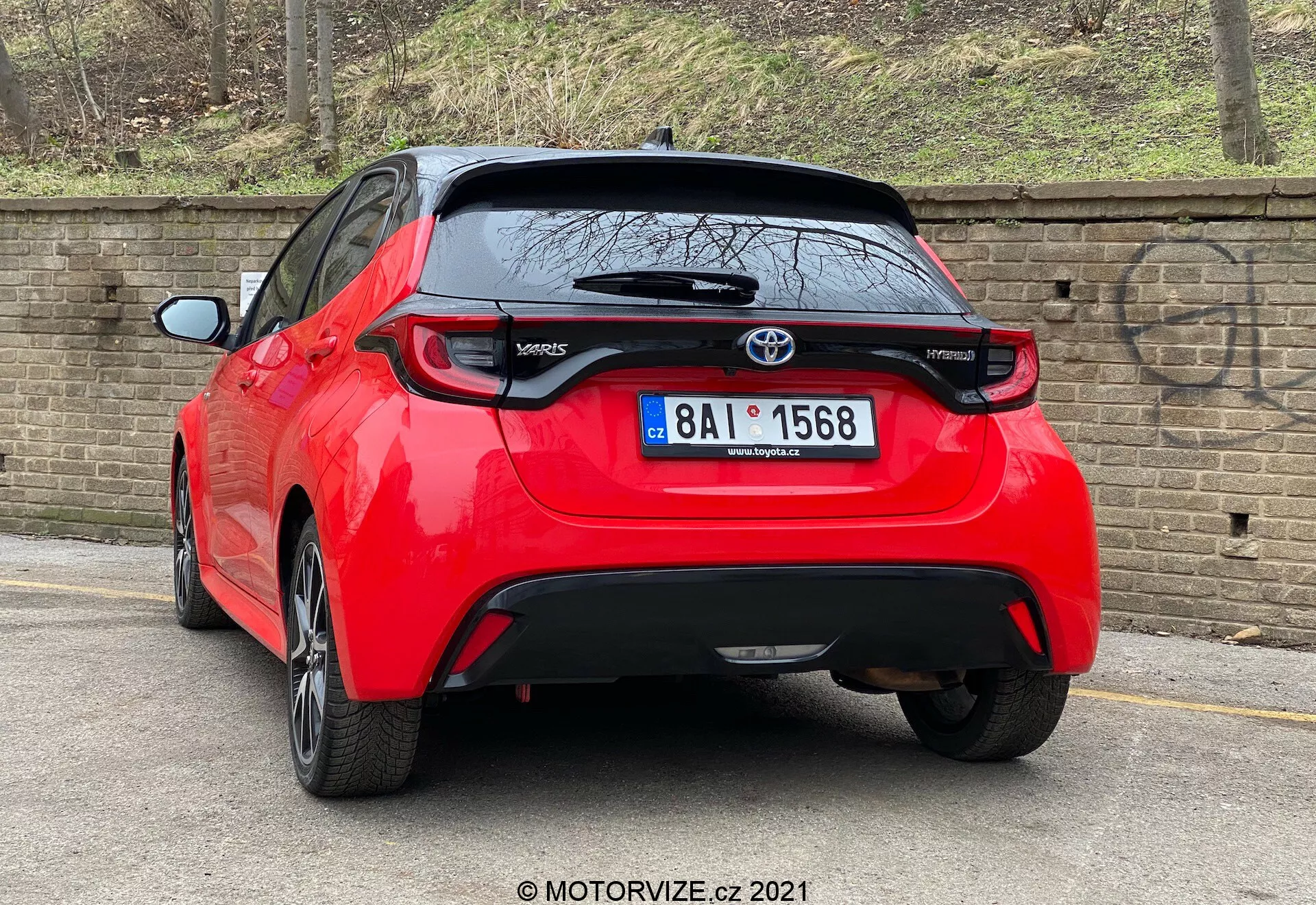 En högupplöst bild av en röd Toyota Yaris 2020 hatchback visad från en bak-trekvarts vinkel. Fordonet har distinkta svepande baklyktor, ett centralt placerat Toyota-emblem ovanför en troligen bakåtriktad kamera och en 'Yaris'-modellsymbol nedan till vänster på bakluckan. Under registreringsskyltområdet finns en svart trimdel som kan vara av plast eller kolfiber, med en liten bakspoiler som sträcker sig över bakrutan. Bilen, parkerad nära en trottoarkant på en asfalterad väg bredvid en tegelvägg, antyder en urban eller bostadsmiljö.