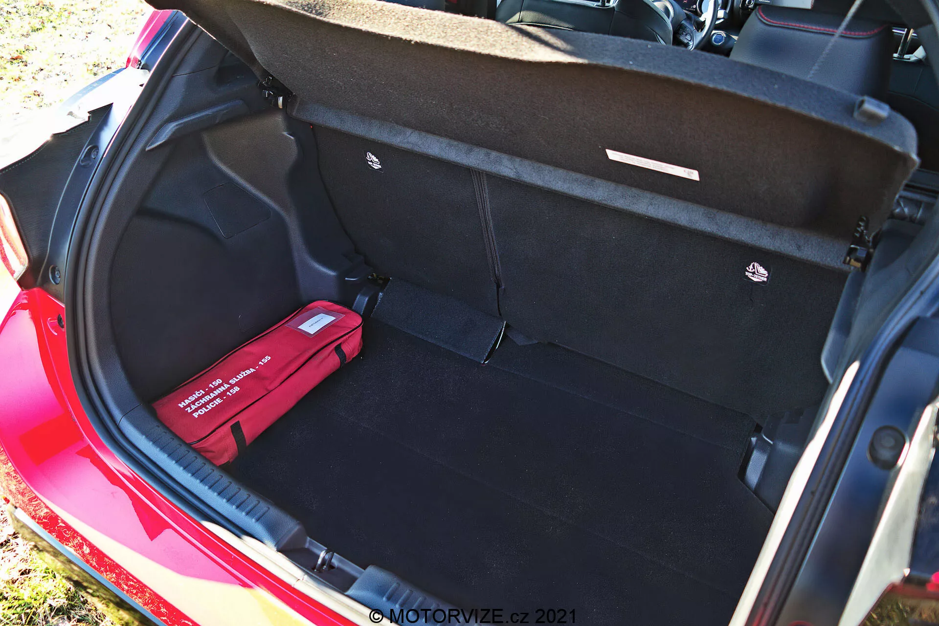 Bagageutrymmet i en röd Toyota Yaris 2020 sett från en högre vinkel, som visar dess rena, rymliga interiör med svart matta, öppen bagagelucka med draghandtag, en plan lastyta, ett lastkroksfäste till höger och en röd första hjälpen- eller nödvägssats till vänster. Denna dagsfotografering framhäver fordonets stora lagringskapacitet och organiseringsfunktioner.