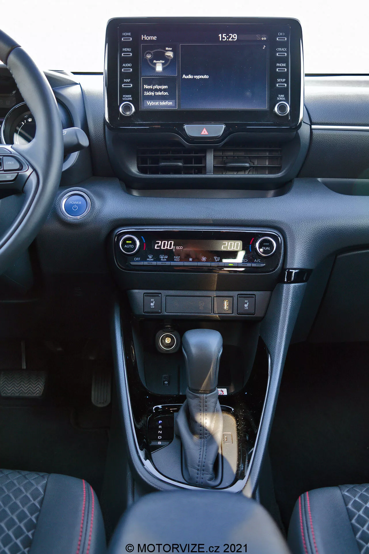 Invändig vy av en 2020 Toyota Yaris med fokus på mittkonsolen och delar av instrumentbrädan. Bilden, tagen från framsätet på passagerarsidan, visar ett digitalt infotainmentsystem med pekskärm som visar en startskärm med tiden 15:29 och olika menyval, centrala luftventiler och användarvänliga kontroller för luftkonditionering och uppvärmning med en digital temperaturdisplay inställd på 20,0 grader. Mittkonsolen har en läderklädd växelspak med en silveraccent och en manuell parkeringsbroms. Interiören är designad med mörka toner och metalliska detaljer, vilket skapar en modern och sofistikerad estetik.