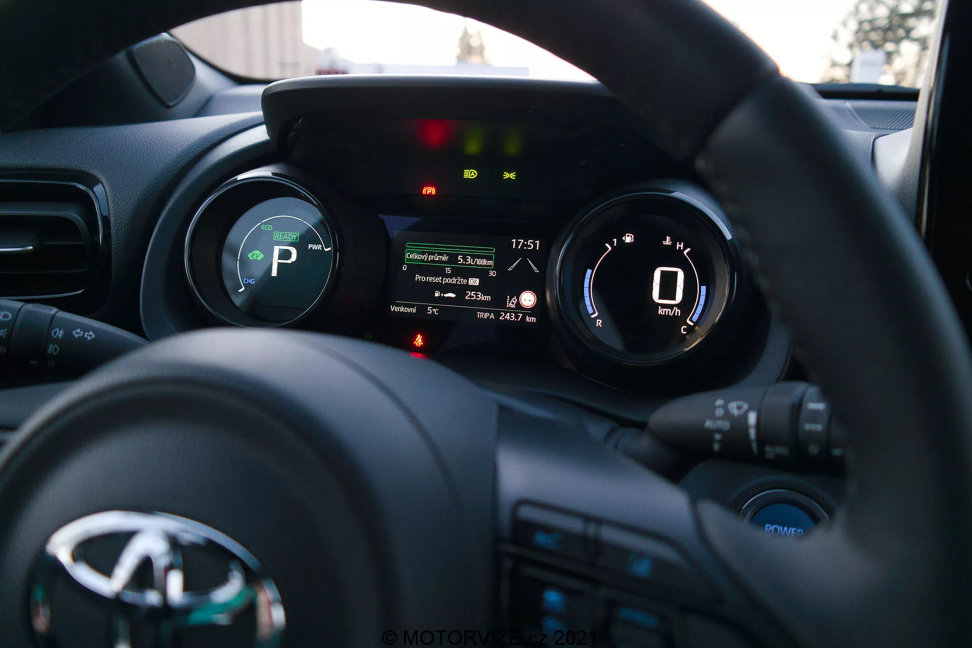 Ett fotografi av interiören på en Toyota Yaris 2020 från förarens perspektiv, med fokus på ratten och instrumentpanelen. Ratten, med tillverkarens logotyp och flera kontrollknappar för media, kommunikation och farthållare, är i botten. Instrumentpanelen bakom ratten har en digital skärm som visar en hastighetsmätare på 0 km/h och indikatorer för växel (P för Parkering), säkerhetsbältespåminnelse, varning för öppen dörr och andra fordonstillståndslampor. Det korta skärpedjupet framhäver instrumentpanelen medan instrumentbrädan och interiören i bilen är mjukt suddiga i bakgrunden. Bilden, troligen tagen sent på eftermiddagen eller tidig kväll, visar modern instrumentering och gränssnittsteknik för fordon.
