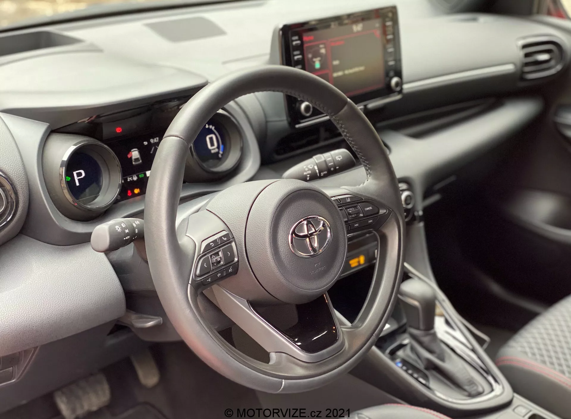 Interiörsbild av en Toyota Yaris 2020 från framsätet på passagerarsidan, med fokus på förarens område. Bilden framhäver en tre-ekrad Toyota ratt med kontrollknappar för ljudsystem, Bluetooth och farthållare, med Toyotas emblem i mitten. Bakom ratten är den digitala instrumentpanelen delvis synlig och visar att bilen är i 'P' (parkering) läge. Mittkonsolen visar infotainmentskärmen och HVAC-kontroller nedanför den, tillsammans med potentiella USB- eller aux-ingångsportar. Bilden visar också en automatisk växelspak med modern design och en läderliknande beklädnad. Interiören har en modern design med en kombination av mjuka material och silverfärgade accenter, tillsammans med mugghållare eller små förvaringsfack intill växelspaken. En delvis synlig dörrpanel avslöjar en kontrollpanel för fönstren, vilket tyder på elektriska fönster.