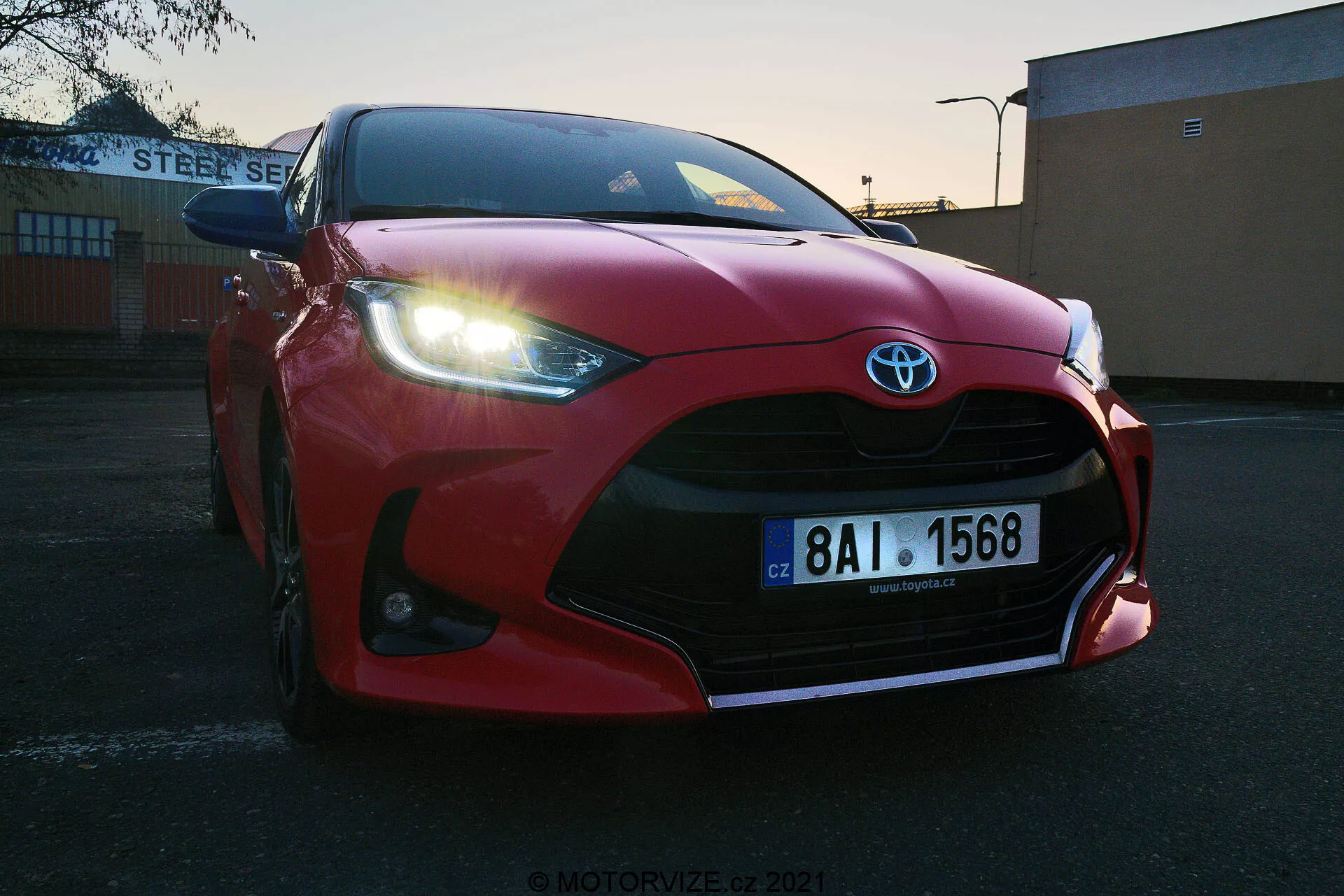 Lågvinkel närbild av röd 2020 Toyota Yaris parkerad utomhus vid soluppgång eller solnedgång. Frampartiet med grill, stötfångare, aerodynamiskt skulpterad huv, skarpa LED-strålkastare och stora luftintag som framhäver Toyota-emblemet. Synlig europeisk registreringsskylt, placerad mot en industriell eller kommersiell bakgrund med 'STEEL SERVICE'-byggnad. Klar himmel med gradientljus som belyser fordonets sportiga design.