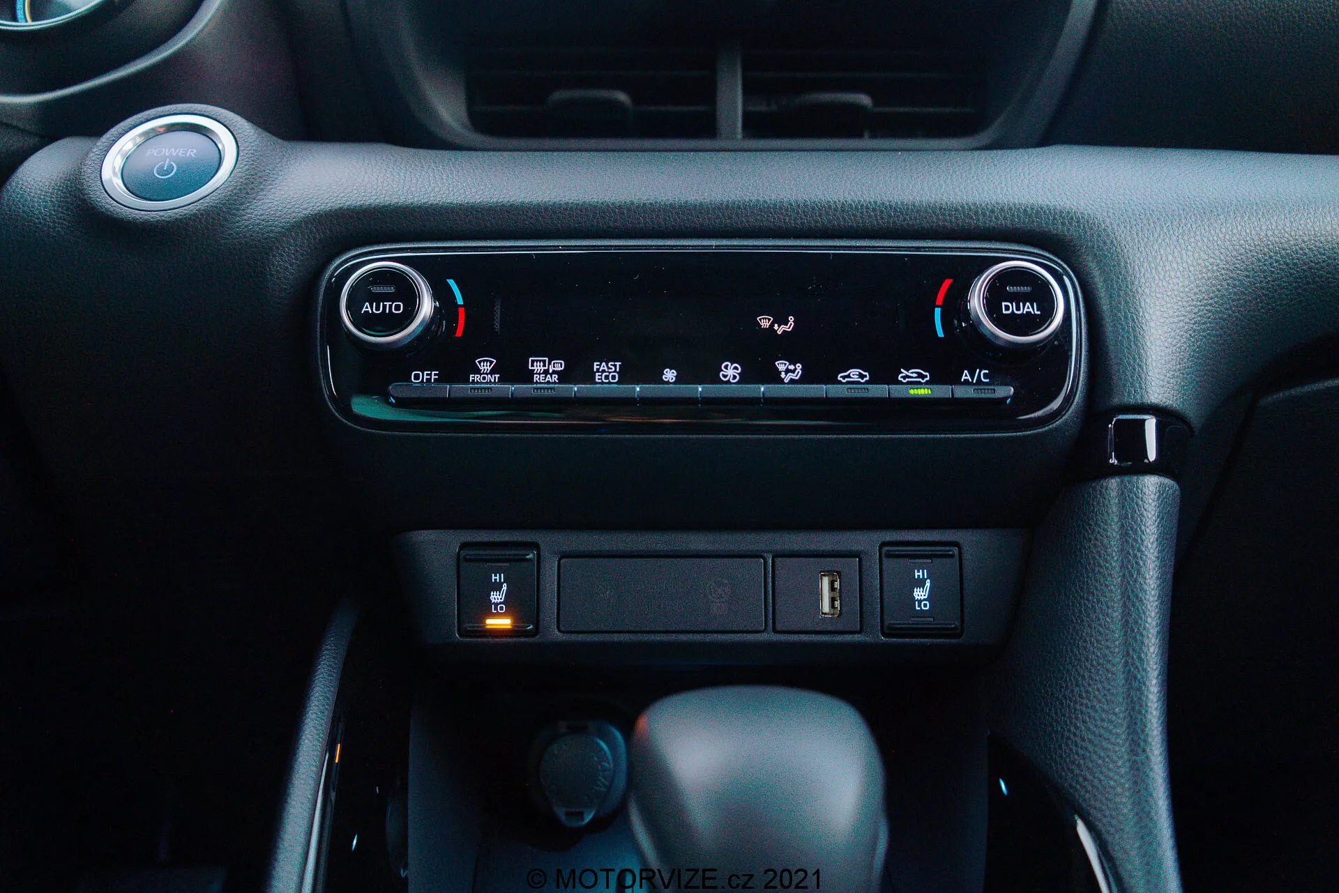 Närbild av instrumentbrädan på Toyota Yaris 2020: Bilden lyfter fram klimatsystemet med tvåzons automatisk klimatkontroll, med 'AUTO'- och 'DUAL'-knappar för oberoende temperaturjusteringar för föraren och passageraren. Under klimatkontrollerna syns ett förvaringsfack med en USB-port och eventuell 12V-strömuttag synliga. Nedanför detta finns tre knoppar: den centrala är växelspaken i 'Park'-läge, vilket indikerar en automatisk växellåda, en mindre vridknapp till vänster sannolikt för körlägesval, och en tryckknapp till höger som kan vara en elektronisk parkeringsbroms. Instrumentbrättets material har ett mörkt färgschema med en premium strukturerad finish, vilket skapar en modern och användarvänlig interiör.