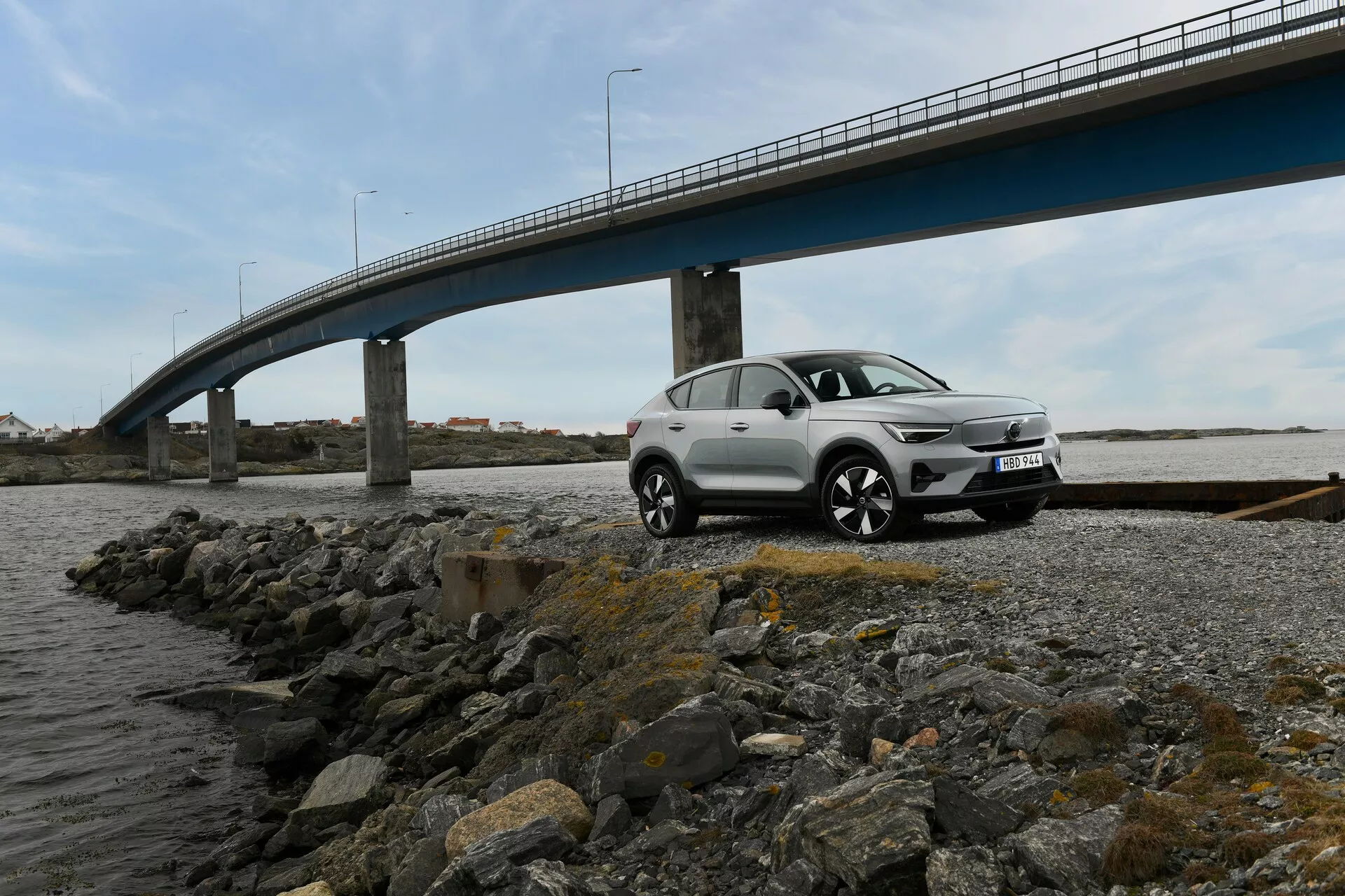 Mellanstora SUV Volvo EC40 1:a generationen 2024 parkerad på en stenig strandlinje med en bro i bakgrunden, visar en tre fjärdedels framsida som framhäver dess frontgrill, strålkastare, stötfångare, motorhuvskontur och sidoprofil mot en molnig dag eller ett mjukt upplyst inställning