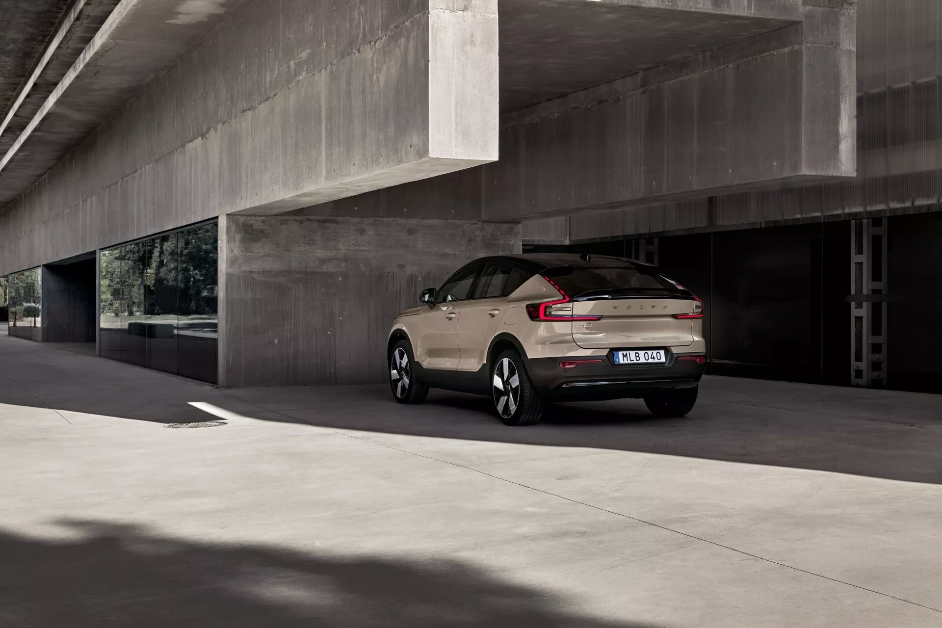 Volvo EC40 2024, 1:a generationen, parkerad under en betongstruktur och visar upp sin moderna SUV-design, med strömlinjeformade bakljus, väldefinierad bakre stötfångare med parkeringssensorer, legerade flerspakiga hjul och en hajfenantenn, i en matt neutral färg, på en mulen dag.