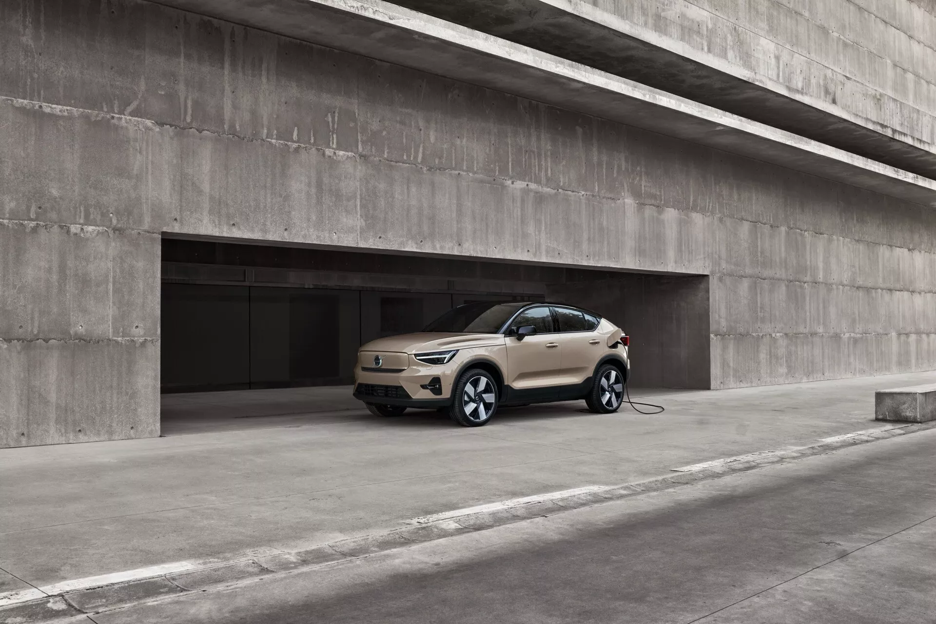 2024 Volvo EC40 1:a generationens elektriska SUV i ljus färg, framvisad i vinkel i en modern urban betongparkering, framhävande dess främre högra kvartal, sidoprofil och delvis bakre vy med distinkta flerspakiga hjul och en synlig laddningskabel ansluten till den främre vänstra skärmen, indikerande dess elektriska eller plug-in-hybrid natur, fotograferad från ett lågt vinkel för att betona markfrigång och hjulstorlek, med en bakgrund som accentuerar dess moderna, strömlinjeformade design med LED-strålkastare och en coupé-liknande lutande taklinje.