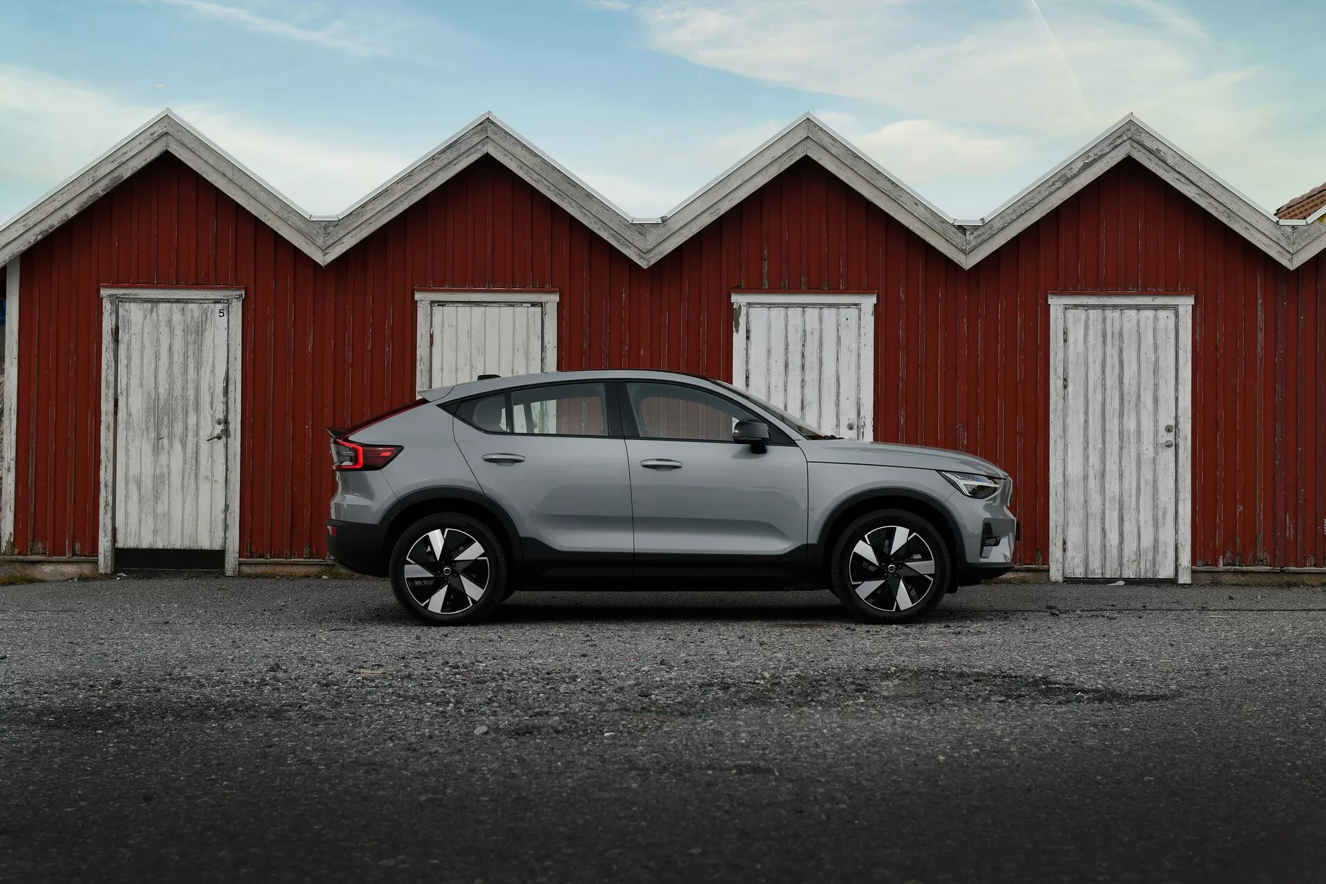 Sidoprofil av den gråa Volvo EC40 Generation 1 2024, visar dess höga markfrigång, eleganta sidobackspeglar, konturlinjer på dörrarna, stiliga aluminiumhjul och kromramade fönster, mot bakgrund av röda trägarage.