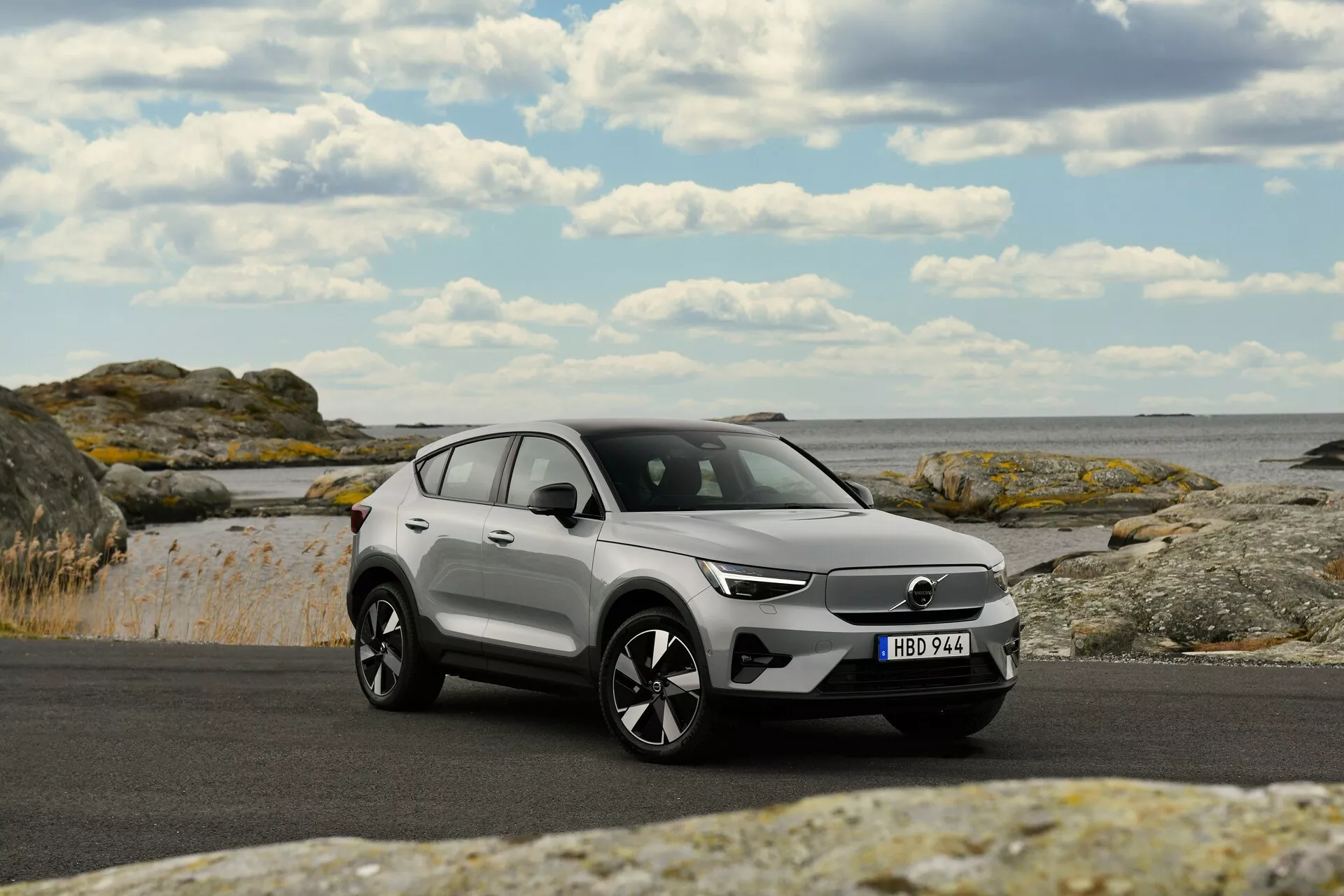 2024 Volvo EC40 1:a generationens moderna SUV parkerad på asfalt med kustbakgrund inklusive klippor och hav under delvis molnig himmel, visar frontgrillen, LED-strålkastare, flerstråliga legeringshjul och tvåfärgat färgschema