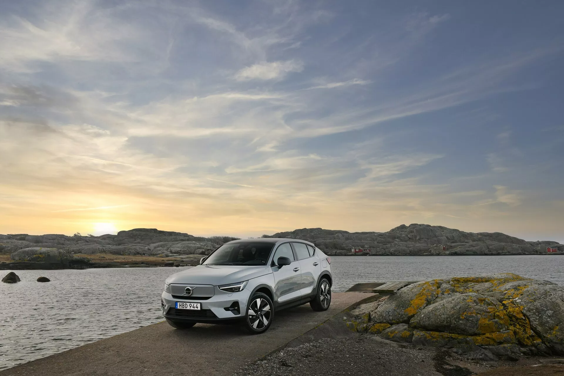 2024 Volvo EC40 1:a generationens moderna crossover/SUV parkerad på ett klippigt kustområde vid solnedgången med synliga LED-strålkastare och avancerade säkerhetsfunktioner, visar fram- och sidoprofiler mot bakgrund av havet och molnig himmel