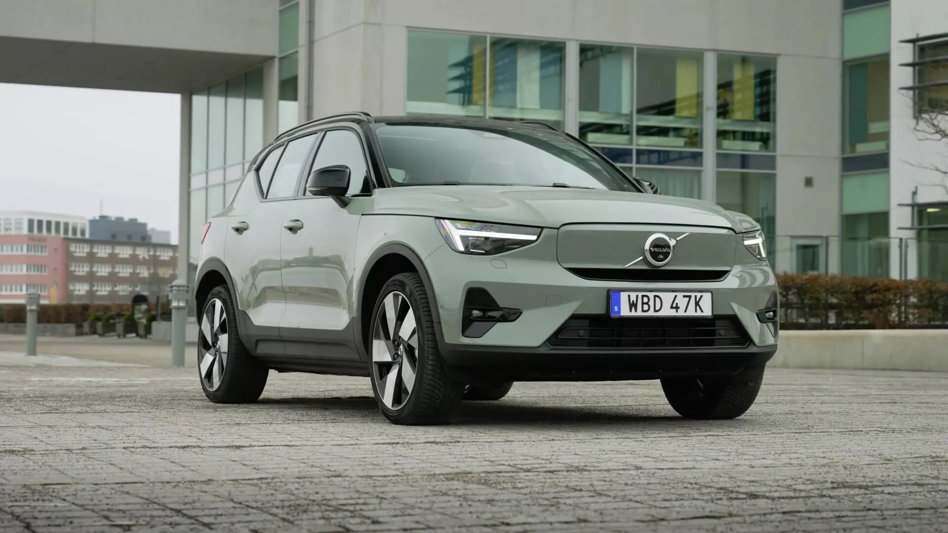 2024 Volvo EX40 1:a generationens elektriska SUV parkerad på ett stenlagt område med stadens byggnader i bakgrunden, som visar dess aerodynamiska frontfasad med stängd grill, eleganta LED-strålkastare, stora flerspakiga lättmetallhjul, modern karossdesign med dörrhandtag i plan med karossen, integrerade blinkers i sidorutorna, fönster med krominramning och avancerad förarassistans-teknik.
