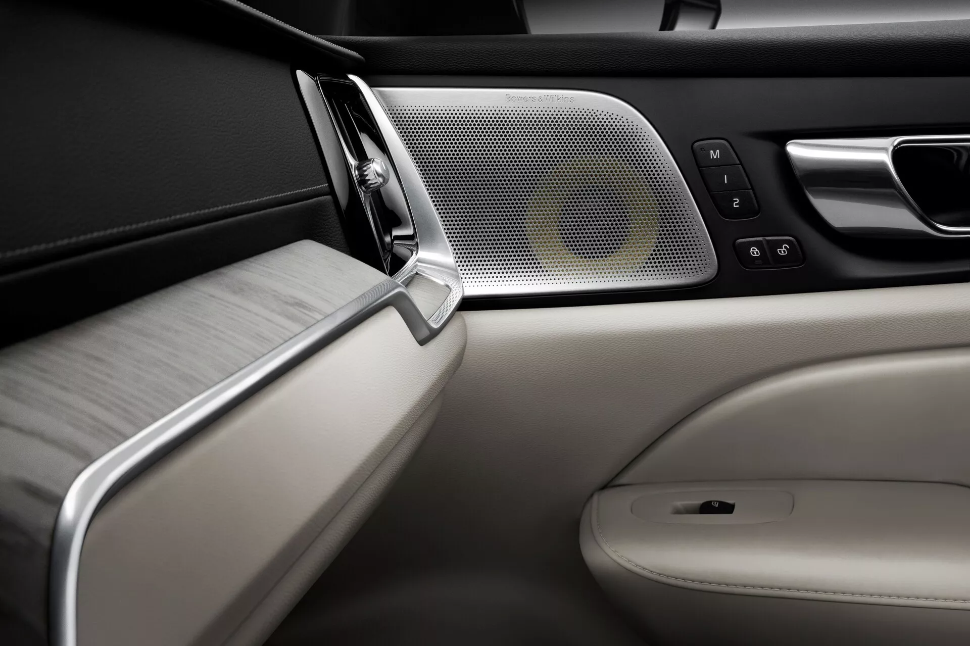 Närbild på passagerarsidans dörrpanel på insidan av en Volvo S60-V60 2018, med fokus på lyxiga designelement. Funktioner inkluderar ett dörrhandtag i metall med krom- eller satinfinish, ett högkvalitativt högtalgaller, ett beige läderklätt armstöd med fönsterkontroller och borstad aluminiumdetaljering, samt en kombination av svart mjukt material och en aluminiumlist i den övre delen. Perspektivet är på ögonhöjd, med betoning på detaljerna i dörrhandtaget och armstödet med ett grunt skärpedjup.
