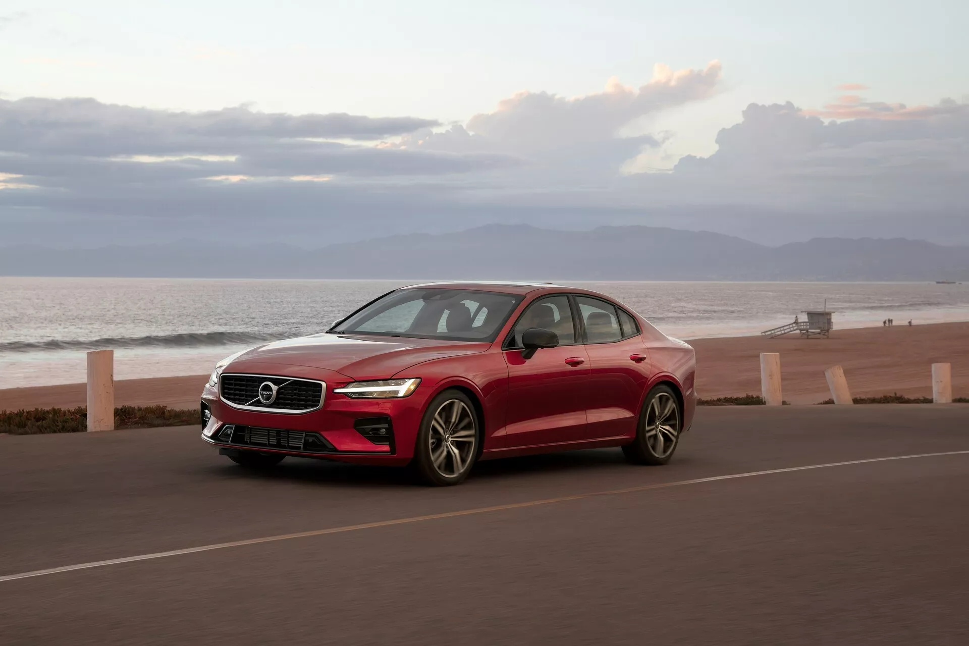 2018 Volvo S60 sedan från trekvartsframvinkel på kustväg, livlig röd, med havet i bakgrunden. Funktioner inkluderar elegant aerodynamisk design, LED-dagljus, flerspårslegeringshjul, karossfärgade sidobackspeglar och soltak. Fångad i tidig morgon- eller kvällsbelysning.