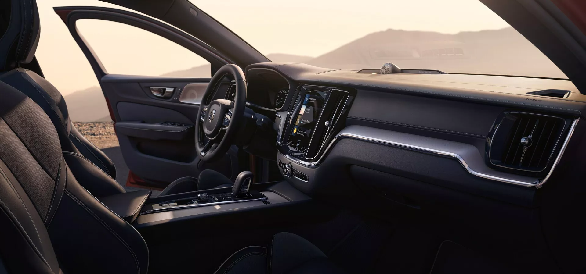 Interiörsbild av en Volvo S60/V60 2018 (3:e generationen) från passagerarens sida. Bilden visar främre kupén, inklusive instrumentbrädan, ratten, mittkonsolen och en del av passagerarsätet. Instrumentbrädan har en stor centralt placerad pekskärm för infotainment och minimala fysiska kontroller, vilket framhäver en stilren, modern design. Ratten är klädd i läder med inbyggda kontroller och sätena är klädda med synliga sömmar. Bakgrunden, synlig genom fönstren, antyder en utomhusmiljö med kullar eller berg.
