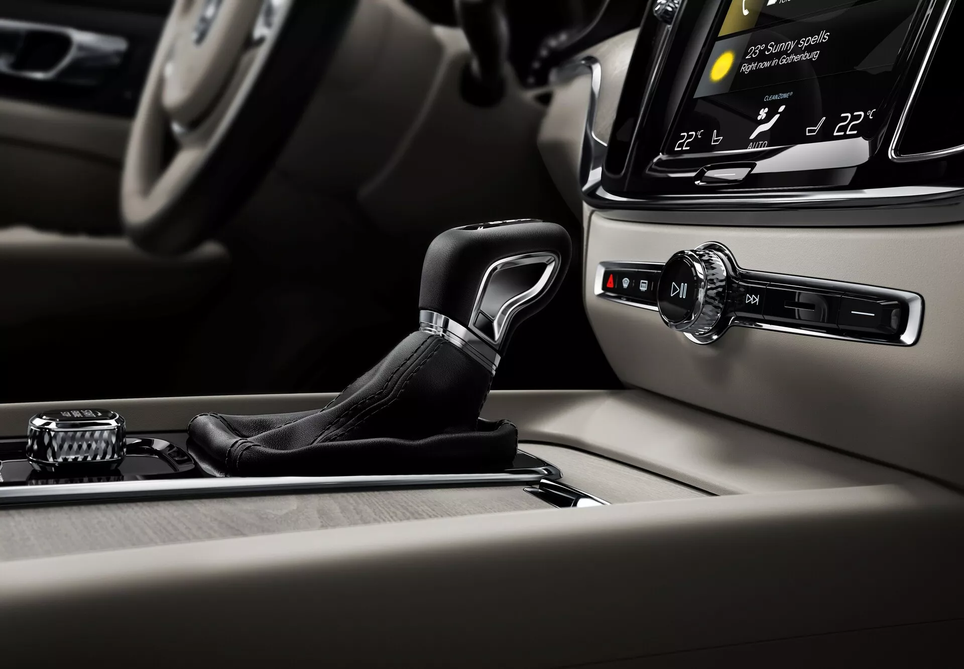 Närbild av interiören i Volvo S60-V60 2018 (3:e generationen), med fokus på mittkonsolen. Den eleganta växelspaken är i vertikalt läge, i park eller neutral, klädd i läder med metalliska inslag. I bakgrunden syns en infotainmentskärm som visar klimatkontroll med två zoner inställda på 22°C, vinklad mot föraren. Till höger om växelspaken finns en start/stopp-knapp, vilket indikerar nyckellös startteknik. Till vänster finns en kontrollknapp med flera knappar runtom, använd för att navigera i infotainmentsystemens menyer. Konsolen är prydd med premiumträ och borstad metall, vilket speglar fordonets lyx och hantverk.