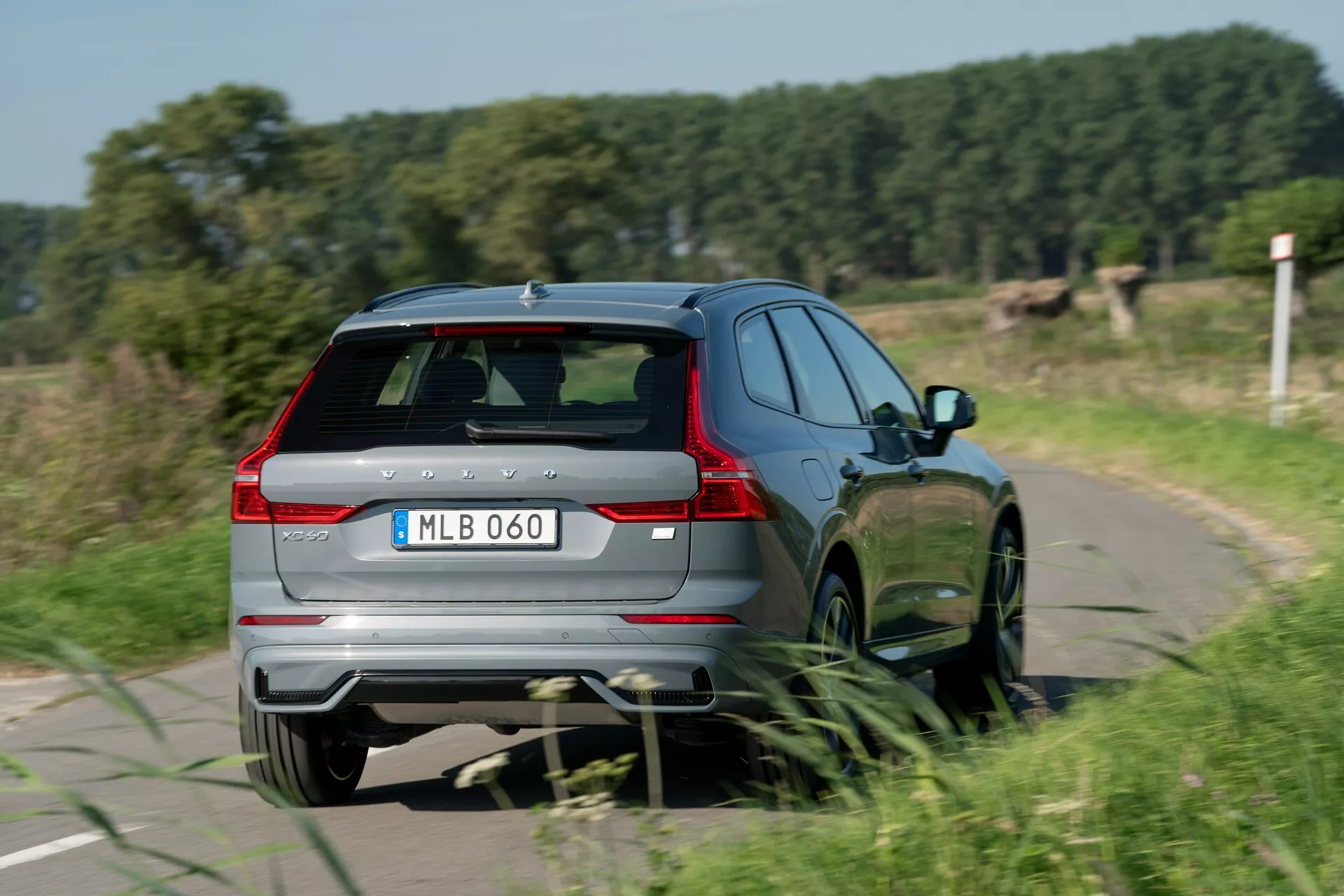 Bakre trekvartsvy av en Volvo XC60 2021 (andra generationens facelift) i rörelse, visar bakre och högra sidans designfunktioner, inklusive en baklucka med bakre vindrutetorkare, spoiler och tredje bromsljus, bakre stötfångare med integrerade reflexer och dubbelt avgasystem, utdragna LED-baklyktor, ren högra sidoprofil med bakre passagerardörr och flerspoke legeringshjul, och modellmärke. Bilen kör på en smal landsväg, vilket fångar en känsla av rörelse med lätt rörelseoskärpa på hjulen.