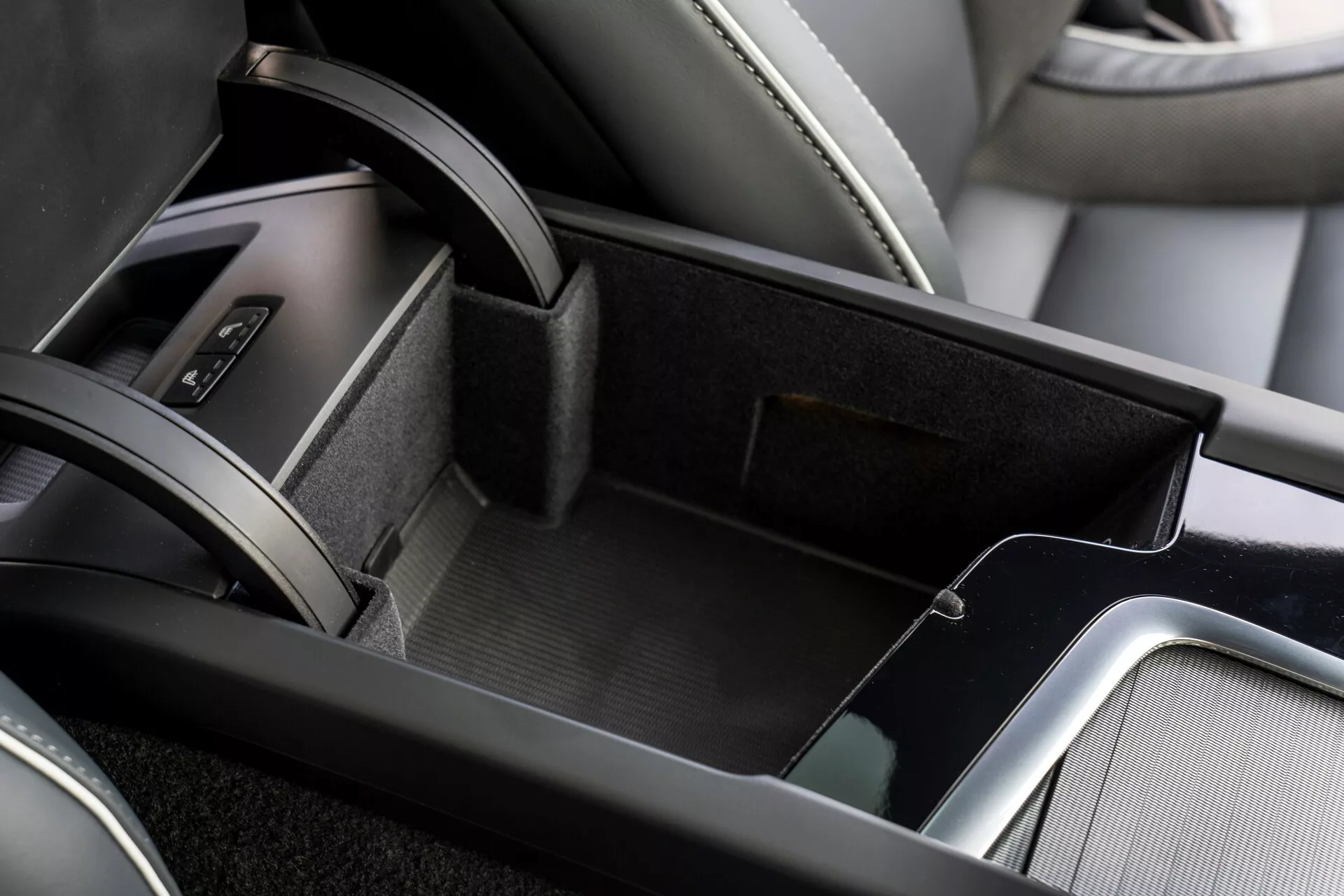 Närbild av mittkonsolområdet i interiören på en Volvo XC60 2021, som visar det öppna förvaringsfacket som är fodrat med filtliknande material, dubbelnivåförvaring med en mindre övre bricka, läderklädd armstöd och metalliska accenter som ger en sofistikerad känsla. En del av växelspaksområdet med lädertäckt damask är synlig i bildens kant.