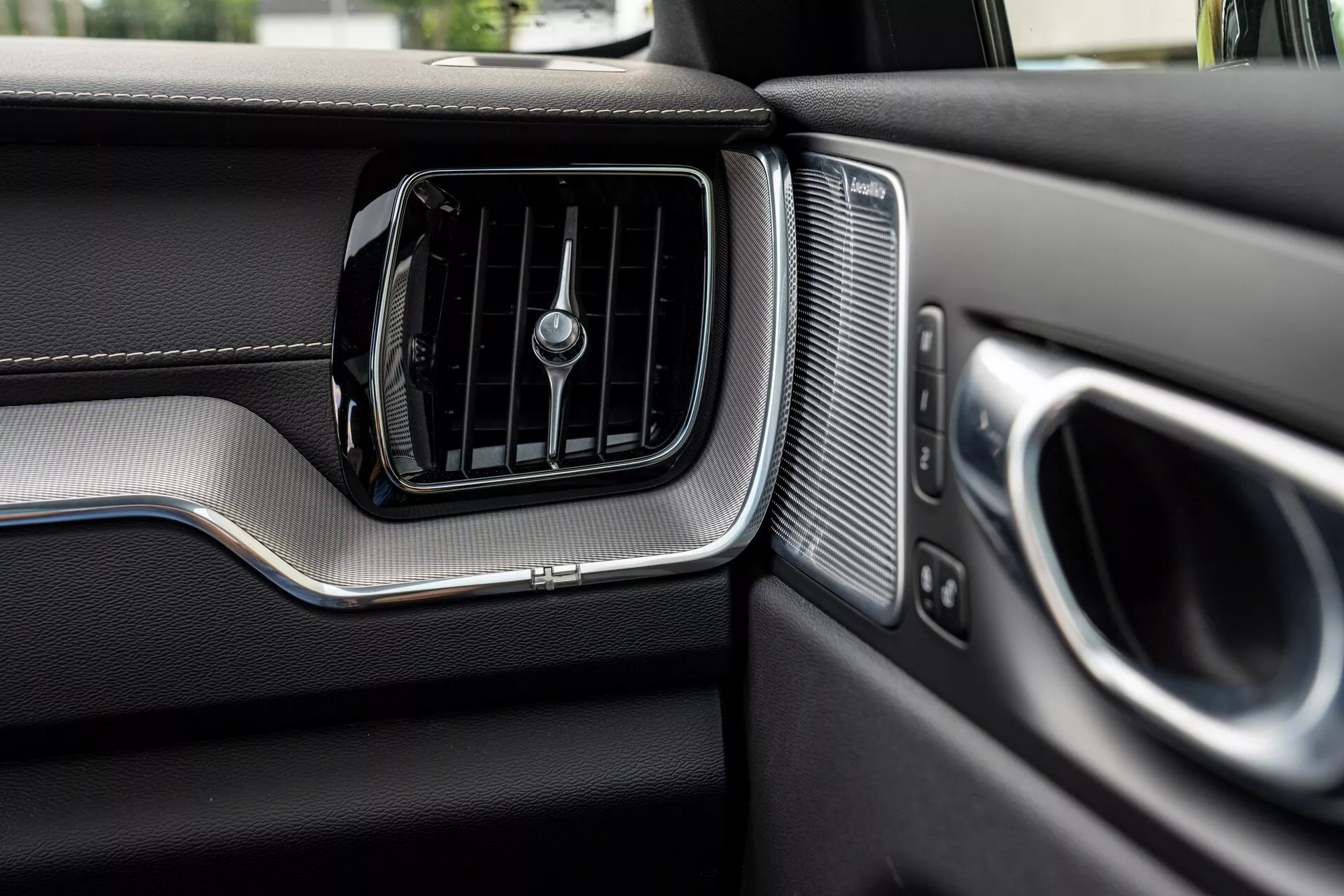Närbild av passagerarsidans instrumentbräda i en Volvo XC60 2021 (2:a generationens facelift). Bilden fokuserar tydligt på luftkonditioneringsventilen med horisontella spjäll och en central rotationsknapp, omgiven av en borstad metallaccent. Till höger om ventilen finns ett högtalargaller med jämna perforeringar, intill den delvis synliga dörrarmstödet och handtaget, även de med metallaccent. Instrumentbrädan visar material som liknar läder på den övre delen och ett annorlunda texturerat nedre område separerat av en metallremsa, vilket betonar fordonets premiumdesign och hantverk.