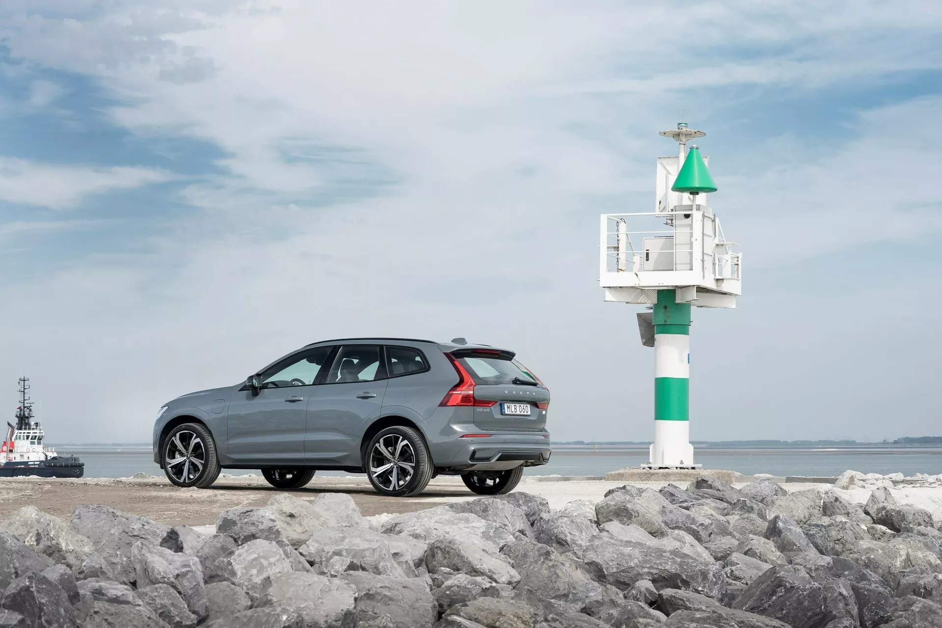 Fotografi av en Volvo XC60 2021 andra generationens ansiktslyftningsmodell, fotograferad ur en trekvart bakre vinkel på en stenig yta, med en maritim miljö i bakgrunden. Den mörkgrå bilen med metallfinsh har en elegant, sportig taklinje, fler-ekrade fälgar i legering, bakre parkeringssensorer och dubbla avgasrör. Till höger om bilen syns en liten fyr eller signal, med en bogserbåt eller lotsbåt i vattnet, vilket förstärker kustatmosfären.