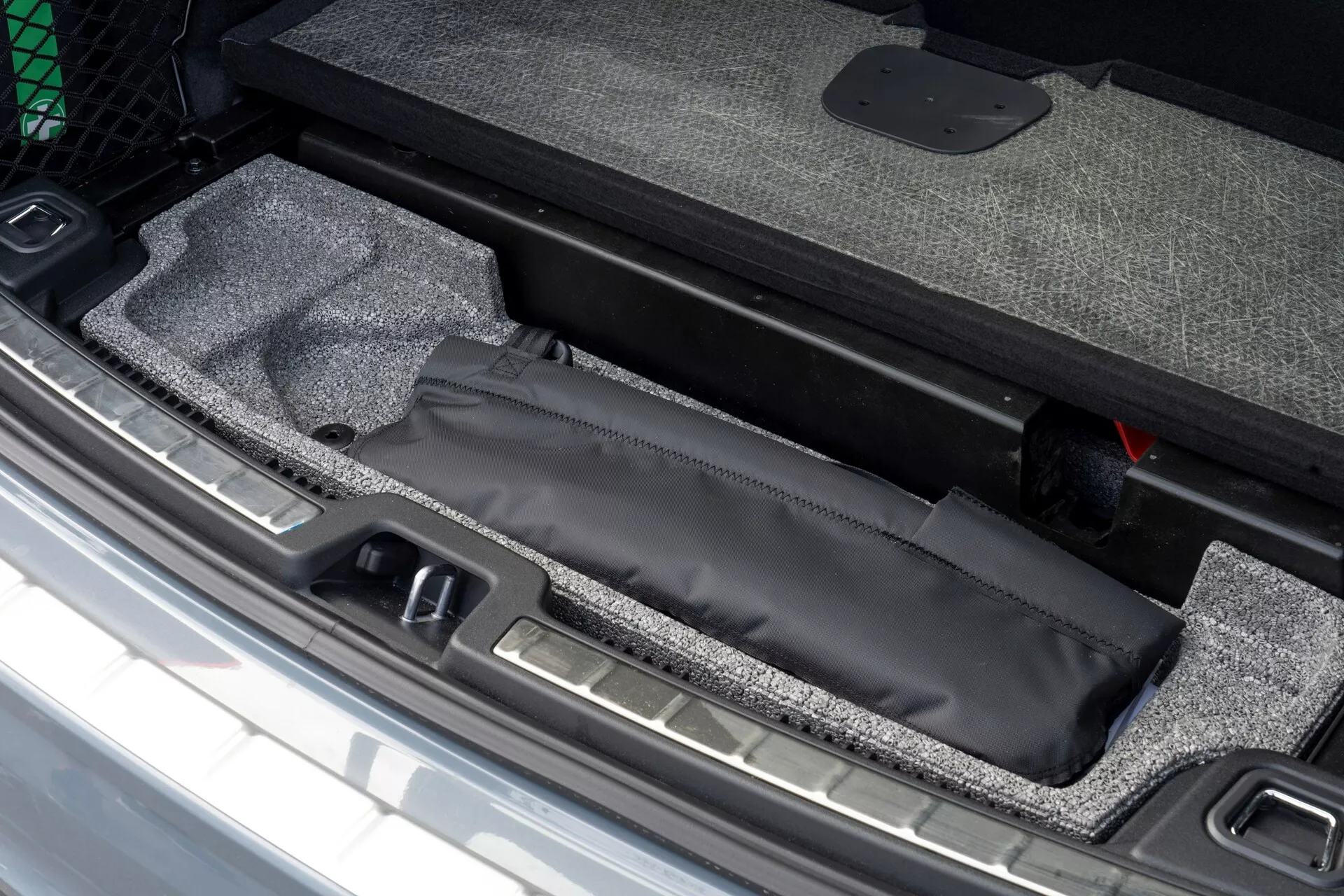 Närbild av Volvo XC60 2021 bagageutrymme som visar dold förvaringsutrymme under bagageutrymmet med svart påse, matta och synlig metalltröskel, bredvid rött nödsituation verktyg.