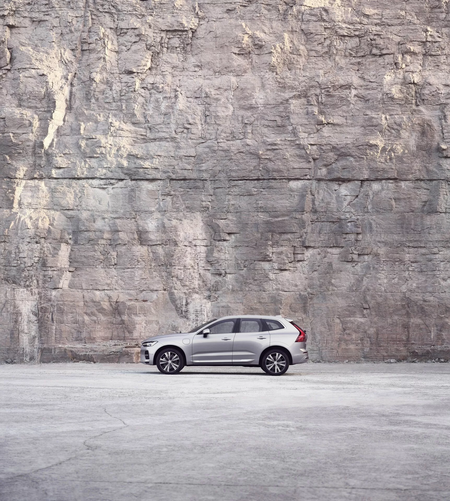 Sidoprofilvy av en Volvo XC60 2021 (2:a generationens facelift) parkerad framför en stor texturerad sten- eller betongvägg, som visar upp sin eleganta och lyxiga design med mörk metallisk färg, flerspoked fälgar, tonade rutor och svarta B-stolpar under mjukt, diffust ljus.