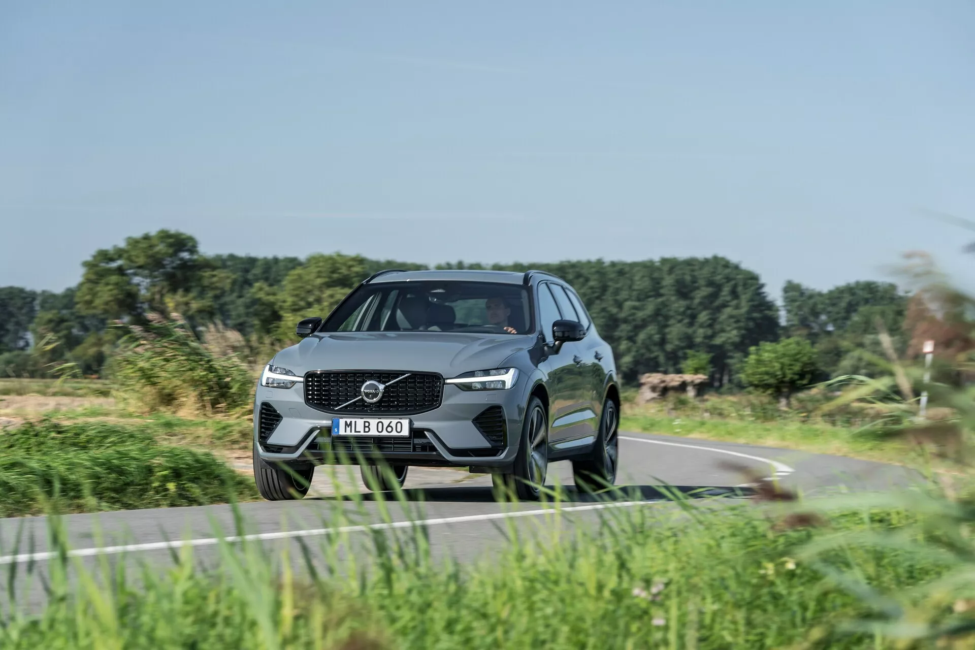 Modern SUV, Volvo XC60 2021 (2:a generationens facelift), fotad från en låg främre trekvartsvinkel som framhäver den dynamiska hållningen och designfunktionerna. Framträdande grilldesign med stort emblem, eleganta LED-strålkastare och skulpterad stötfångare med integrerade luftintag. Aerodynamiskt, lyxigt utseende med konturerade linjer, hög markfrigång, stora flerspoked aluminiomhjul, lågprofil däck, sidospeglar med integrerade blinkers, sluttande taklinje och tonade fönster. Indikerar avancerade tekniska och säkerhetsfunktioner, kör på en landsväg med suddig vegetation som tyder på rörelse.