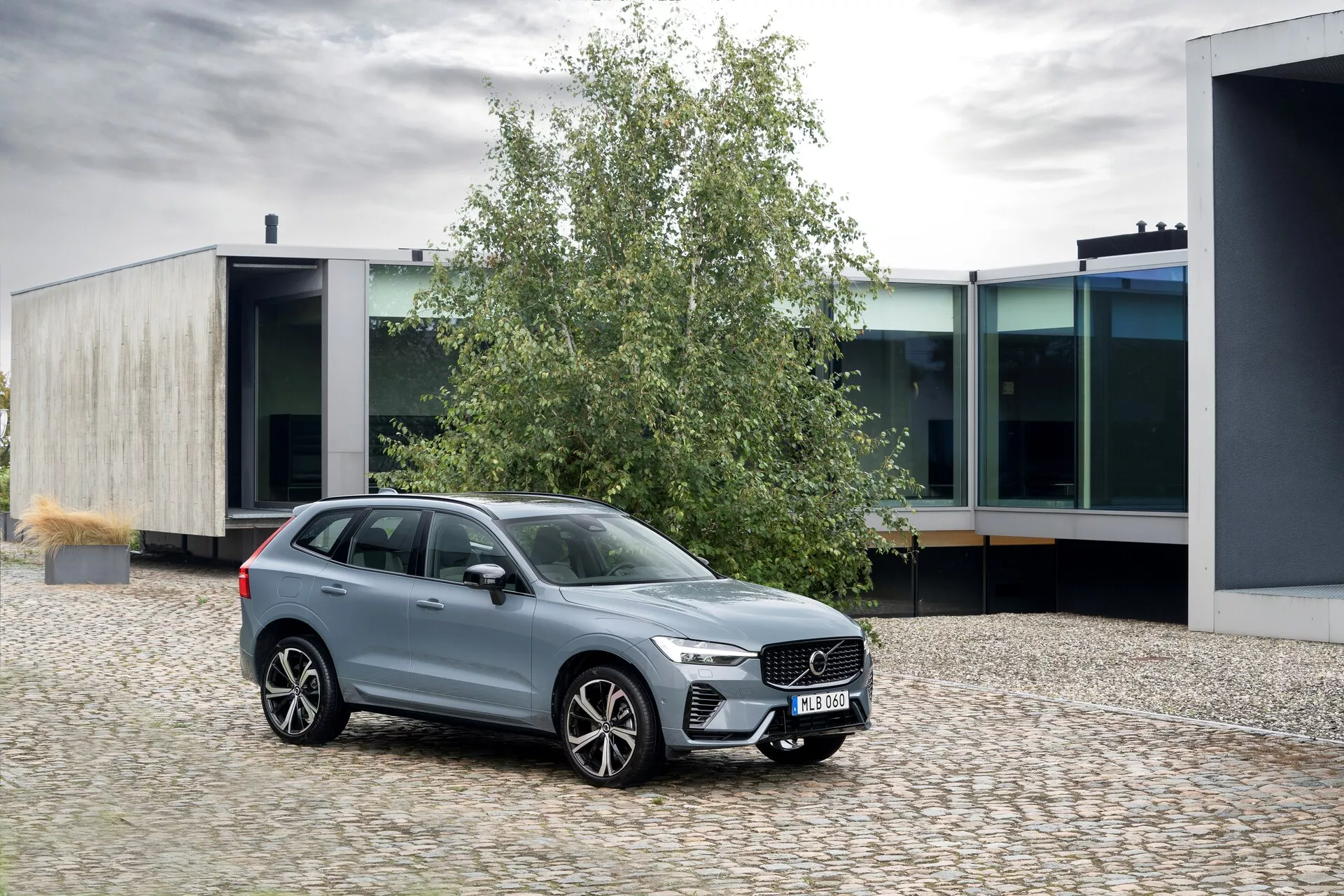 Bild av en Volvo XC60 2021 (generation 2-facelift) parkerad på en kullerstensyta, placerad i en lätt vinkel åt vänster. Från trekvartsperspektiv framifrån syns den framträdande grillen med vertikala ribbor, de eleganta LED-strålkastarna med dagsljus och de stora flerexlade lättmetallfälgarna. Fönstren är omgivna av krom, vilket förstärker den polerade metalliska finishen. SUV:n har en ökad markfrigång, takräcken och lackerade sidospeglar med integrerade blinkers. Miljön är urban med modern arkitektur i bakgrunden, som visar bilens stiliga design och lyxiga egenskaper.
