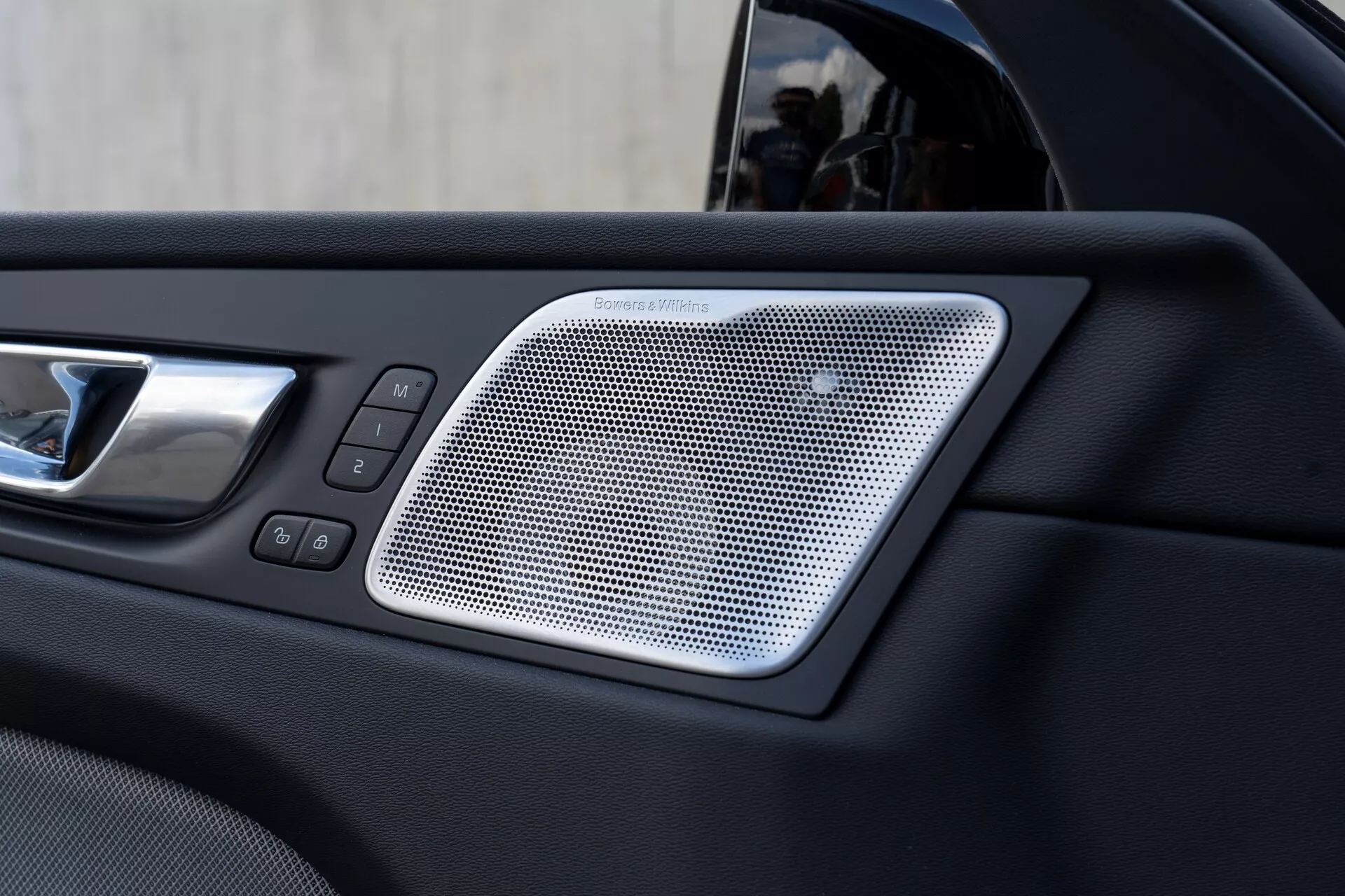 Närbild av den inre delen av en Volvo XC60 2021 (2:a generationen, facelift) bildörr som visar ett Harman/Kardon-högtalargaller med nätmönster, en kromad dörrhandtag och kontroller för el-fönster och dörrlås, vilket betonar bilens lyx och modernitet.