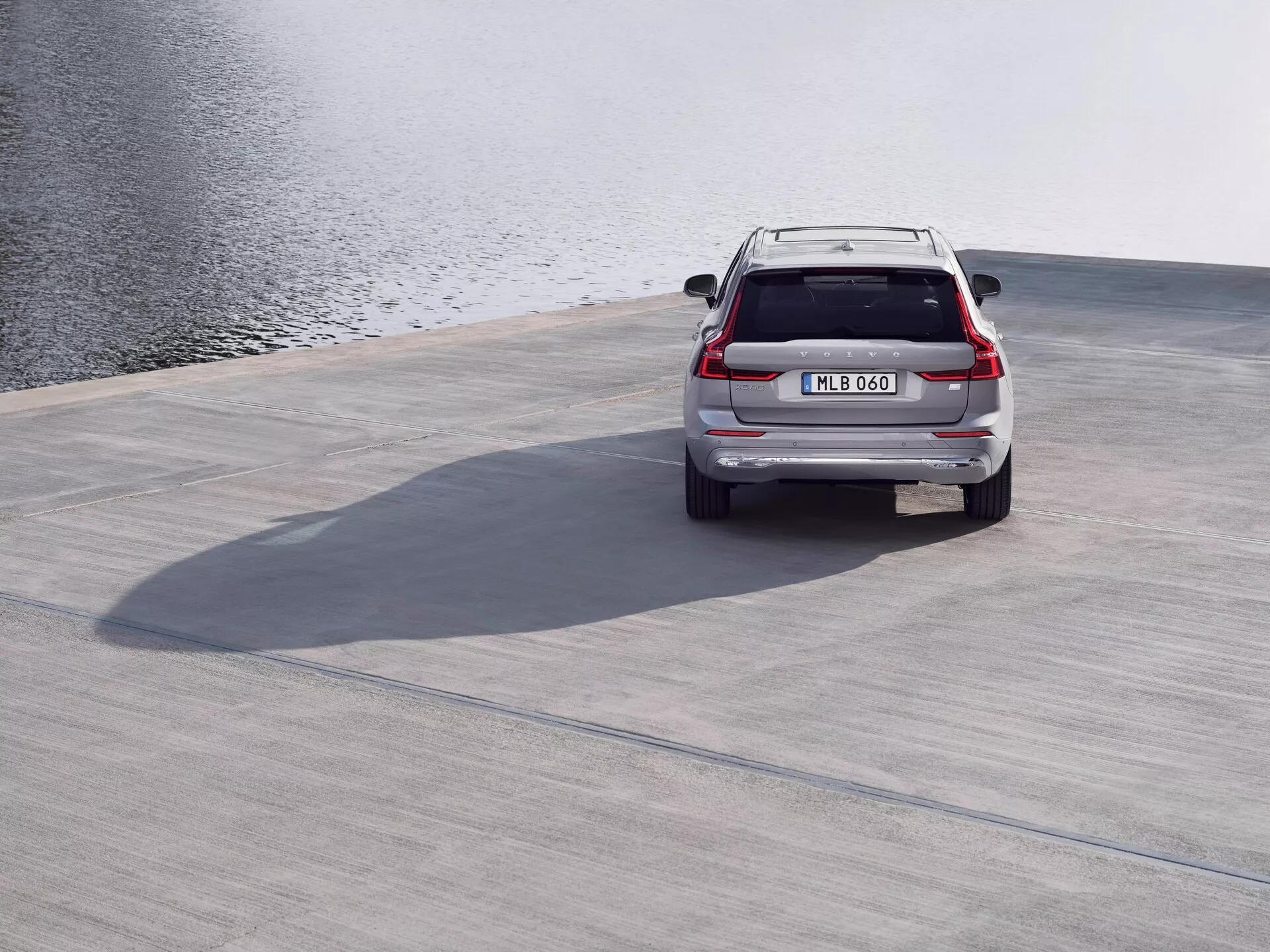 Bakifrån på en silverfärgad 2021 Volvo XC60 (andra generationen, facelift) SUV parkerad på en betongyta nära en vattenspegel, som visar takspoiler, LED-bakljus, baklucka, bakre stötfångare och avgasrör från en hög vinkel med skuggor kastade, under lugna och lätta vindförhållanden.
