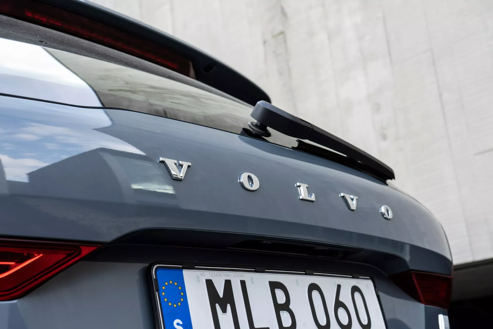 Närbild av bakdelen på en Volvo XC60 2021 (andra generationens facelift). Bilden visar djärva, individuella silverbokstäver som stavar 'Volvo' över bakluckan, en del av bakrutan och en integrerad bakspoiler med en LED-tredje bromsljus. Bilen är mörkgrå eller grafitfärgad, med ett vertikalt LED-bakljus på vänstra sidan som har en 'Tors hammare' design. Under varumärkesnamnet finns en europeisk stilregistreringsskylt med EU-flagga, vilket indikerar EU-registrering och en kromkantad ram. Kameravinkeln är något till höger, med fokus på bokstäverna och bilens karosskanter, med en neutral vit vägg som bakgrund.