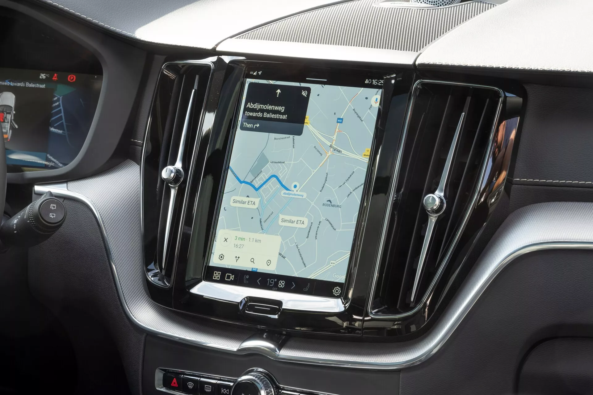 Närbild av mittkonsolen i en Volvo XC60 2021 (2:a generationen, facelift), med fokus på infotainmentsystemet. Fotot, taget från framsätesperspektivet, visar en stor vertikal pekskärm som visar en navigeringskarta. Skärmen är inramad av en metallkant och omgiven av en textur som liknar kolfiber, vilket framhäver moderna och sportiga inredningselement. Fysiska knappar är synliga på sidan av skärmen och nedanför för klimatanläggningen, vilket indikerar en kombination av touch- och fysisk funktion. Delvis syn av ratten på vänster kant bekräftar en vänsterstyrd konfiguration.